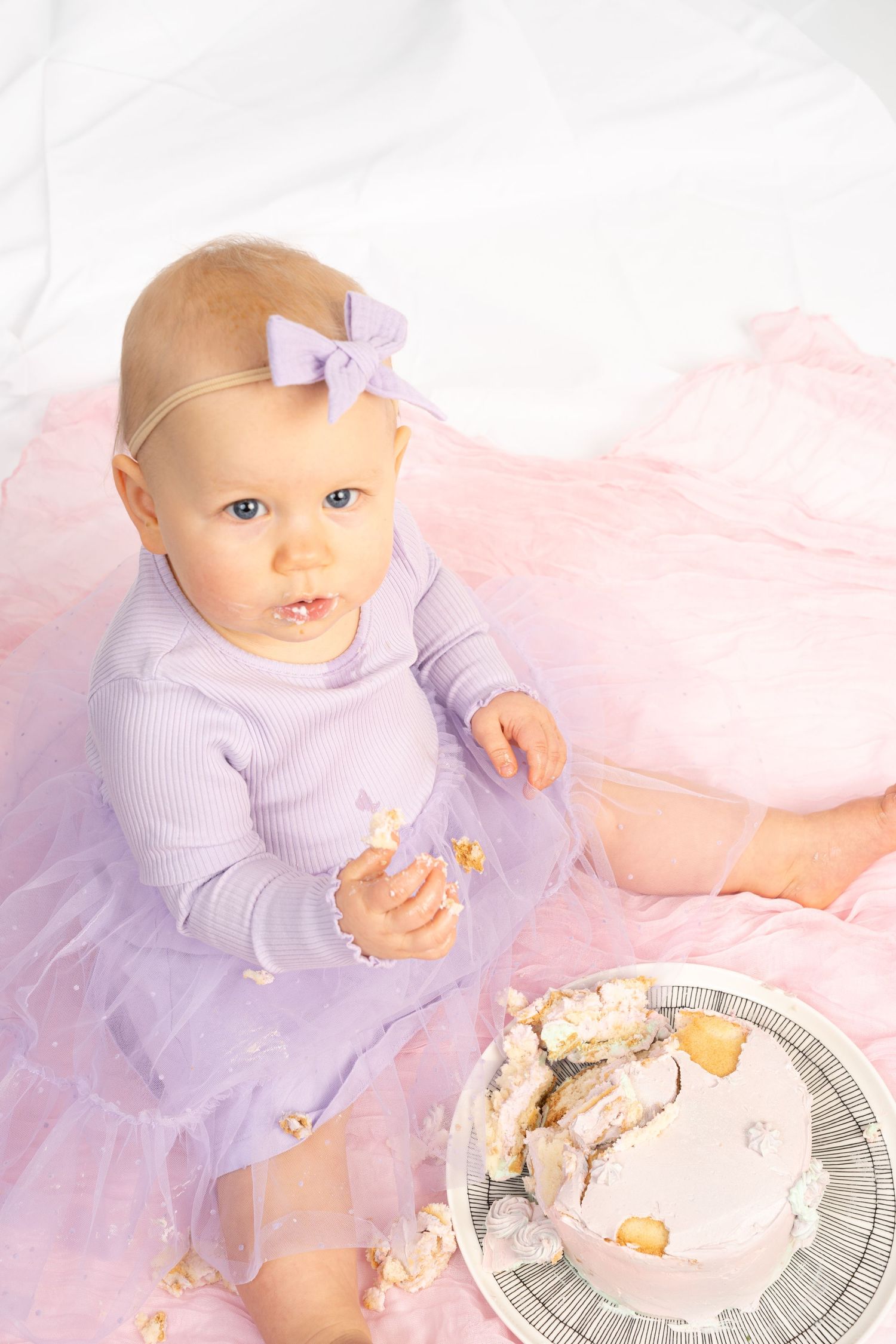 Baby girl in purple tutu dress with bow headband sits beside smashed first birthday cake on pink blanket.