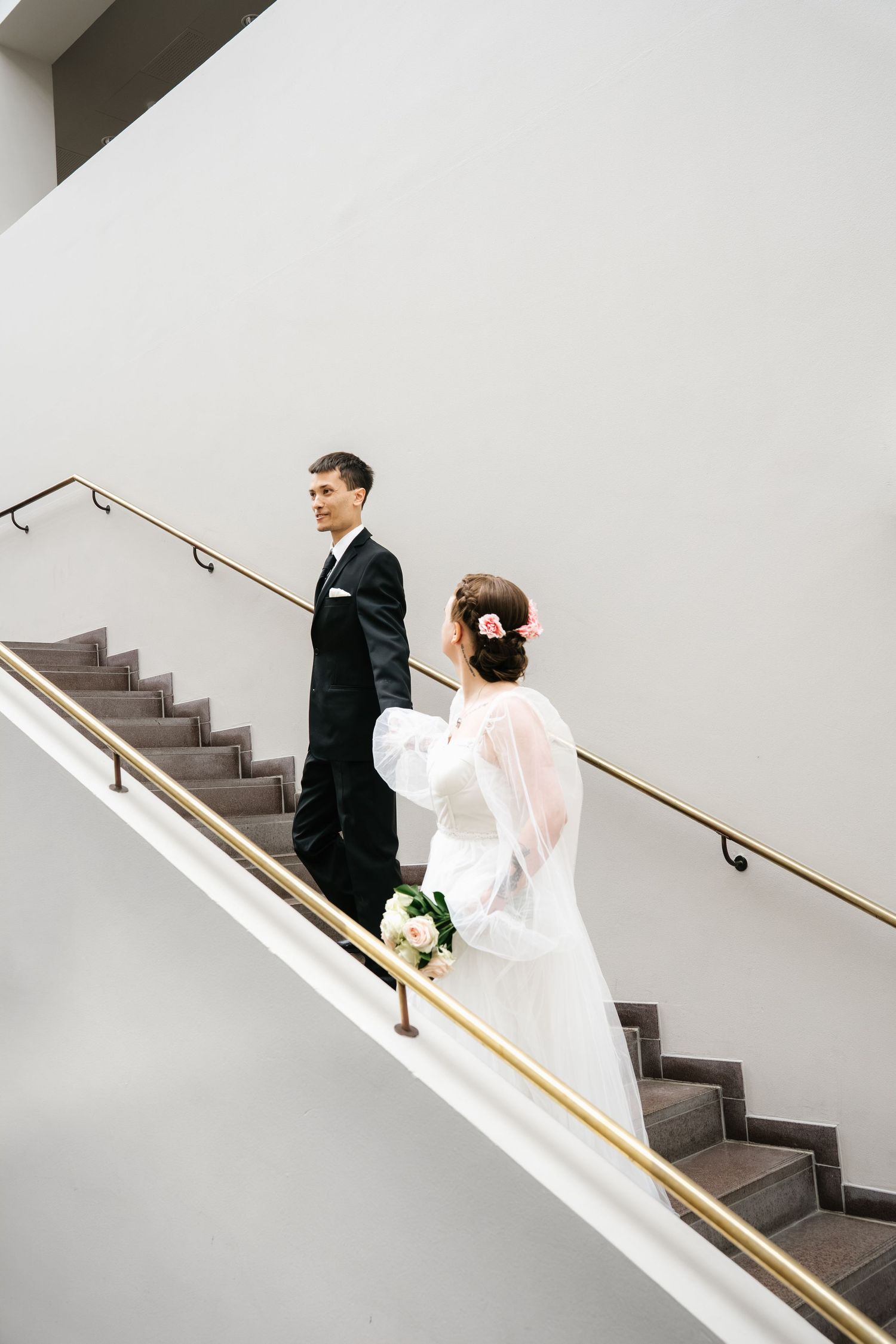Bride and groom walking up elegant staircase, bride holding white bouquet in wedding dress, groom in black suit.