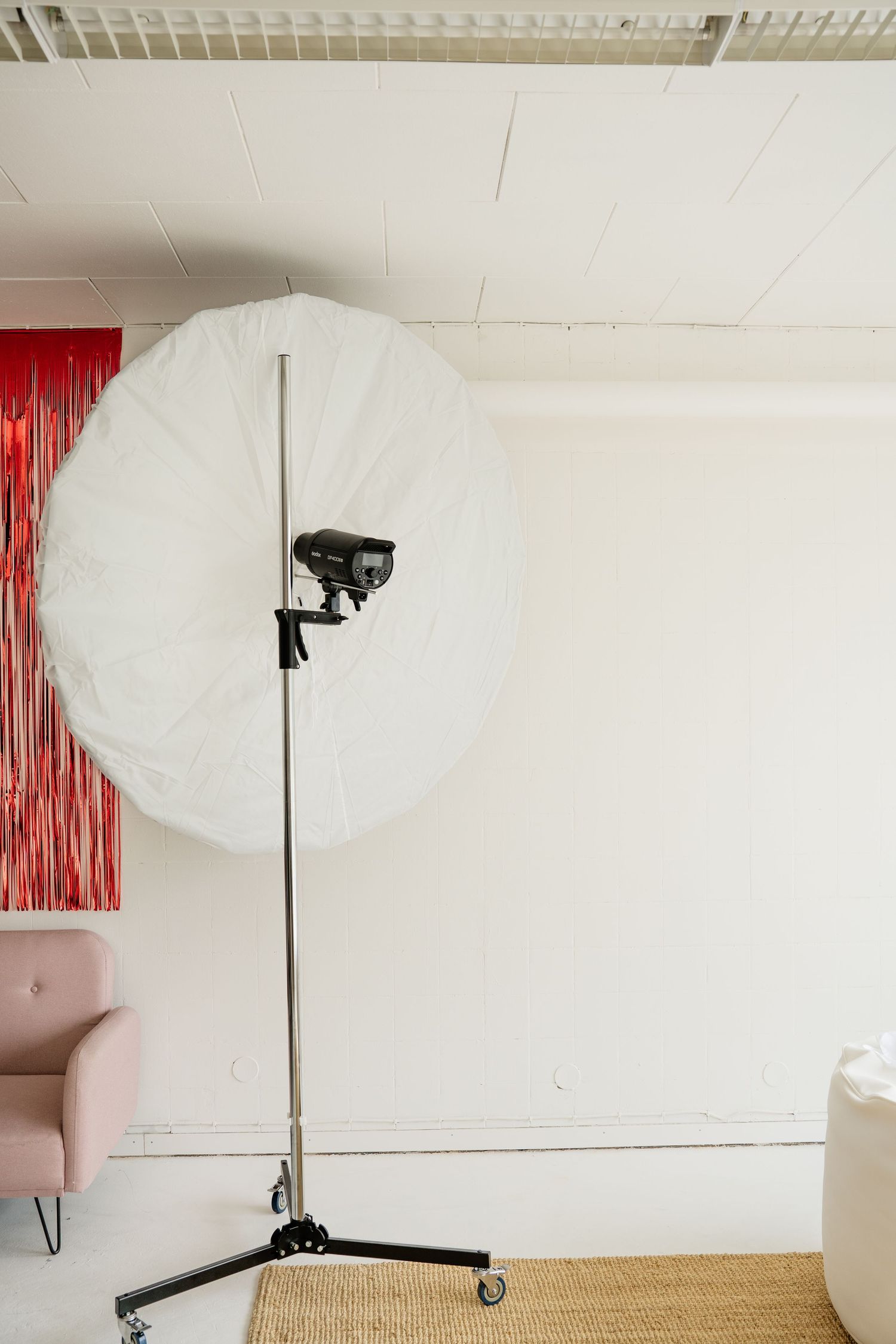 Professional photography studio with large white umbrella light on stand against white wall with pink chair nearby.