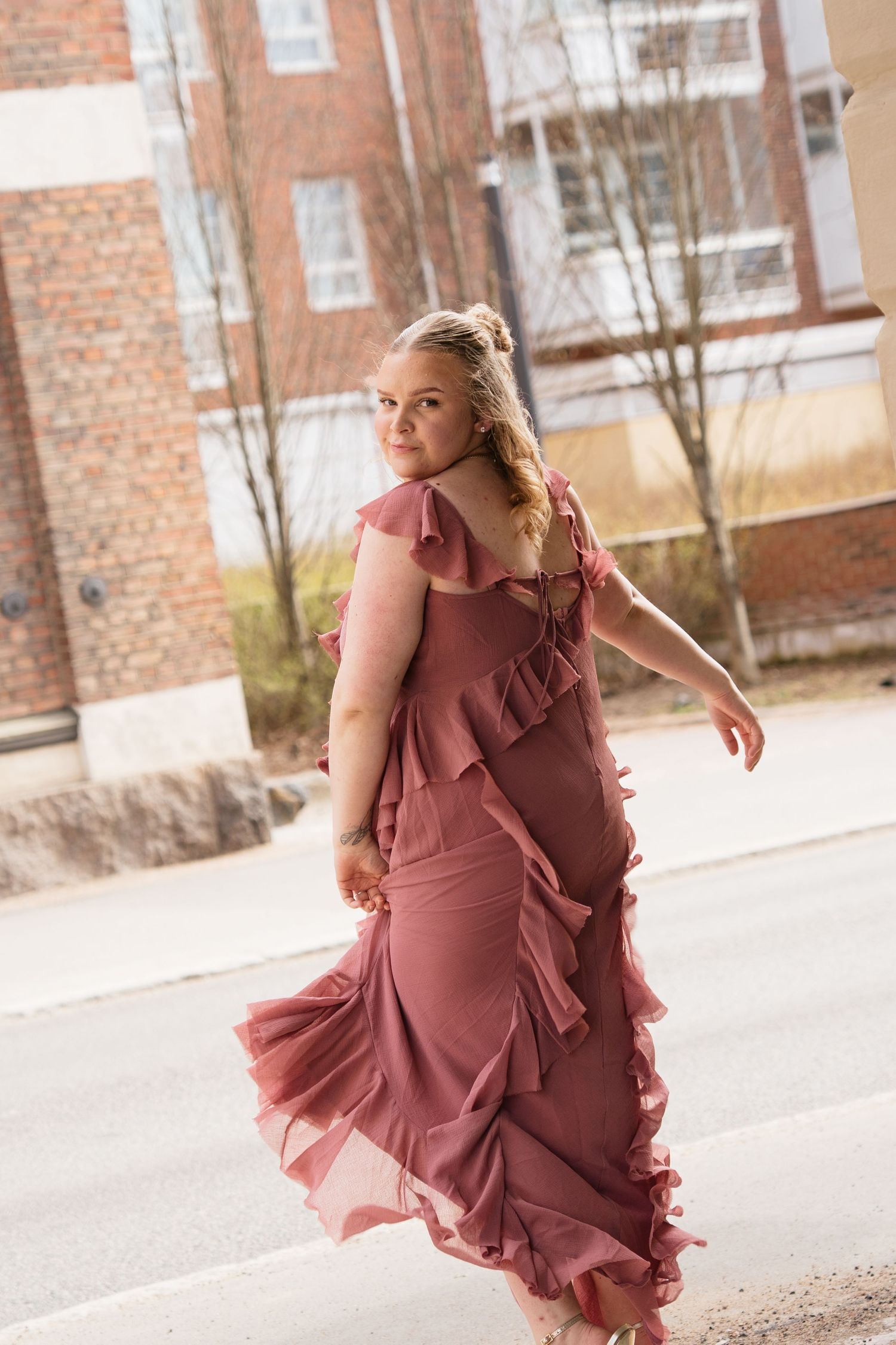 A woman in a flowing pink ruffled maxi dress twirls outdoors on a street, glancing back over her shoulder.