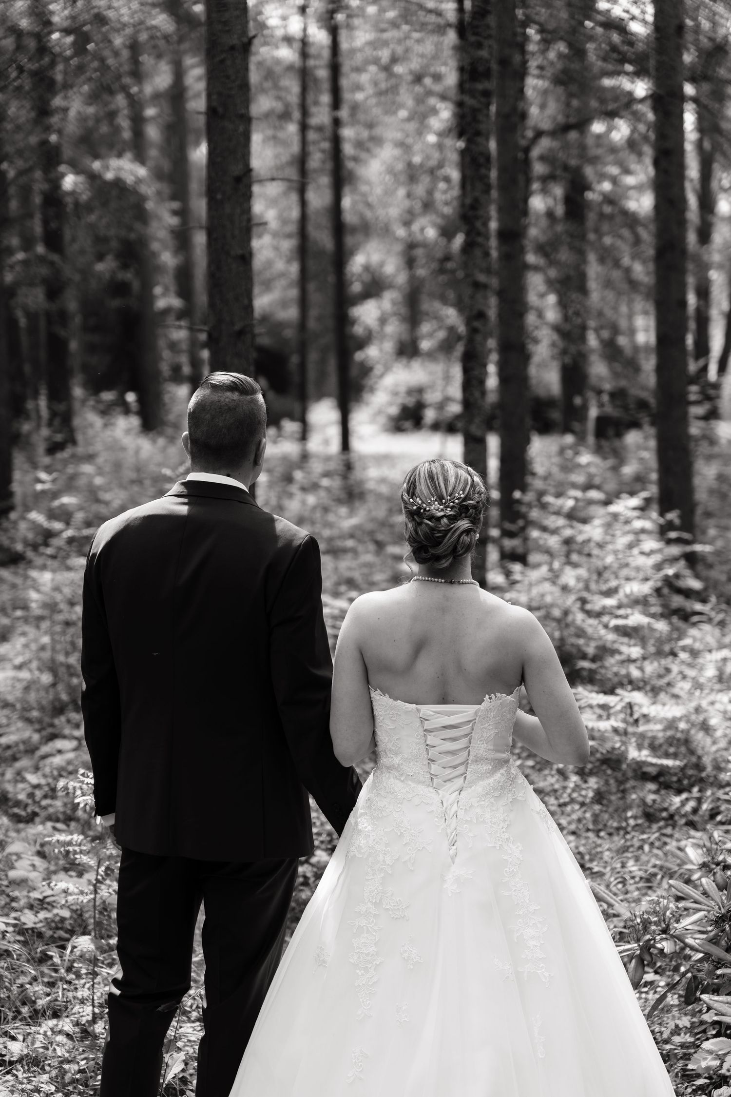 Black and white photo of a bride and groom walking hand in hand through a lush forest on their wedding day.