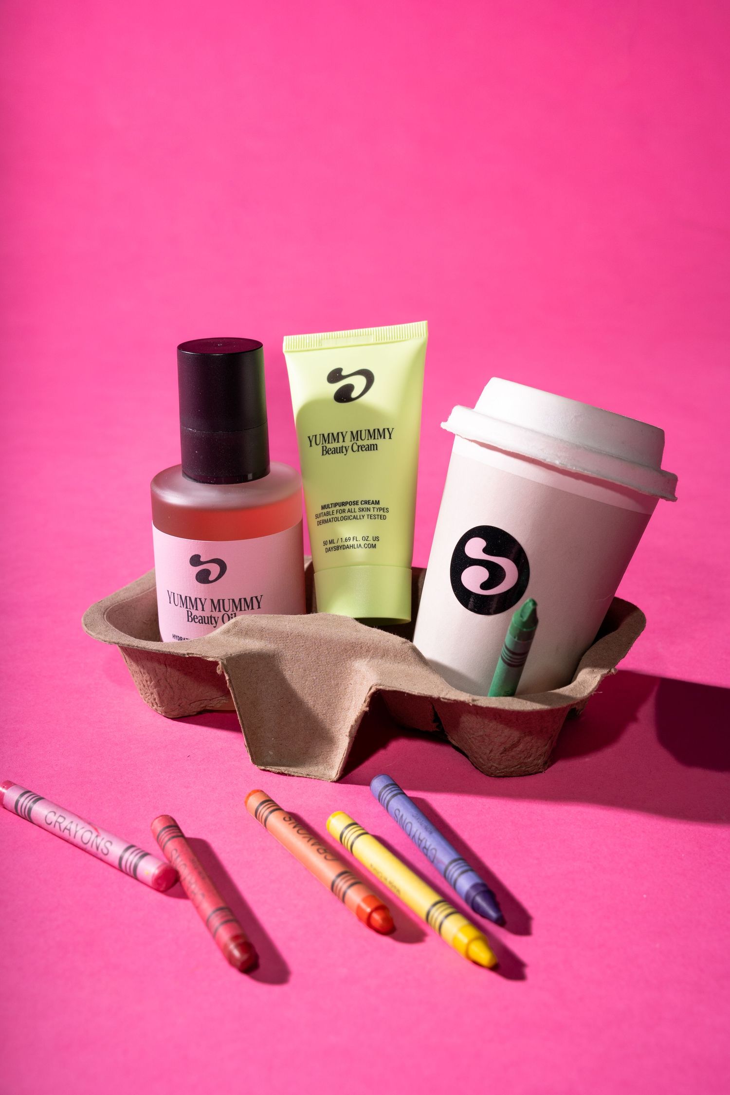 Yummy Mummy beauty products with crayons in a cardboard cup holder on a pink background.