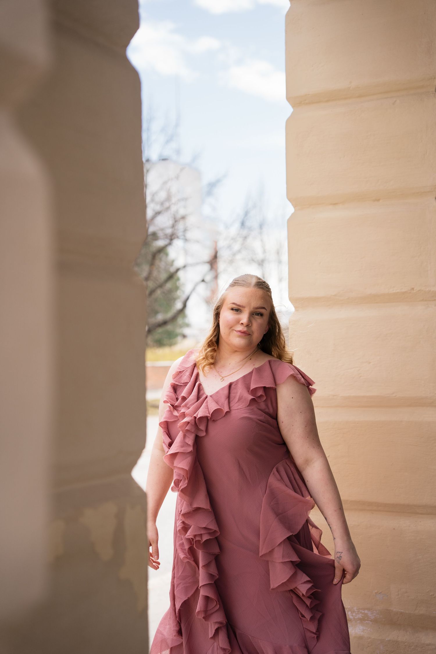 Young woman in pink ruffled maxi dress posing between stone columns outdoors.