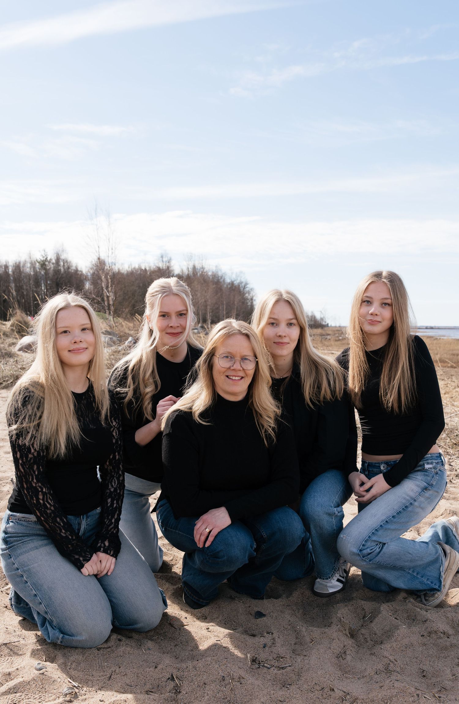 Five blonde women in matching black sweaters and jeans pose together on a sandy beach on a sunny day.
