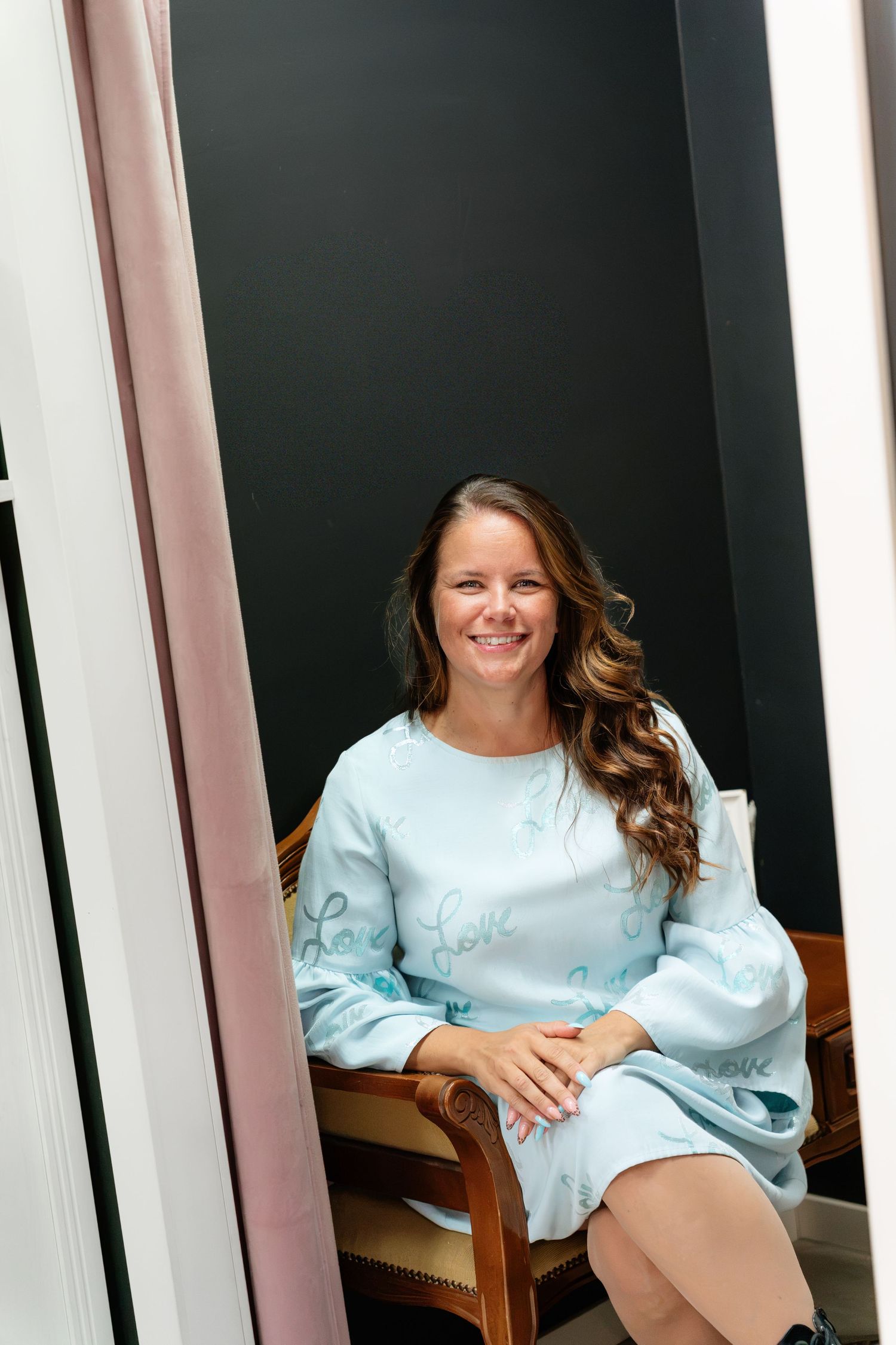 Woman in light blue dress with cursive text sits smiling in a chair against a dark wall.