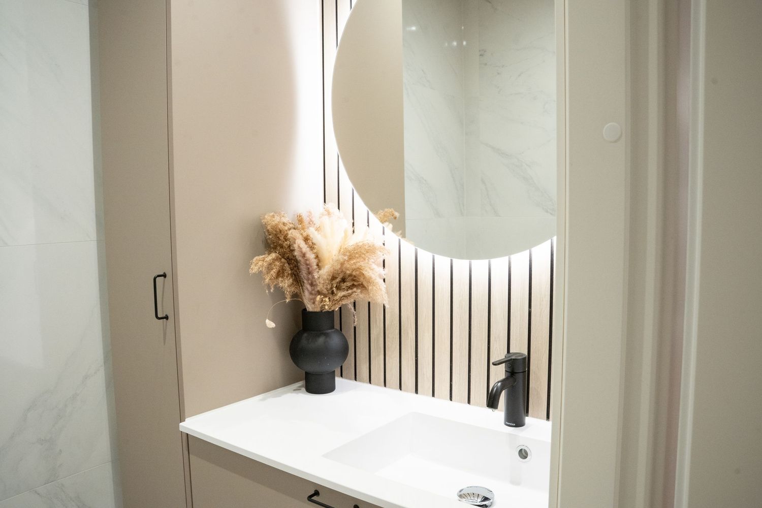 Modern bathroom vanity with round LED mirror, white sink, black faucet, and decorative dried pampas grass in black vase.