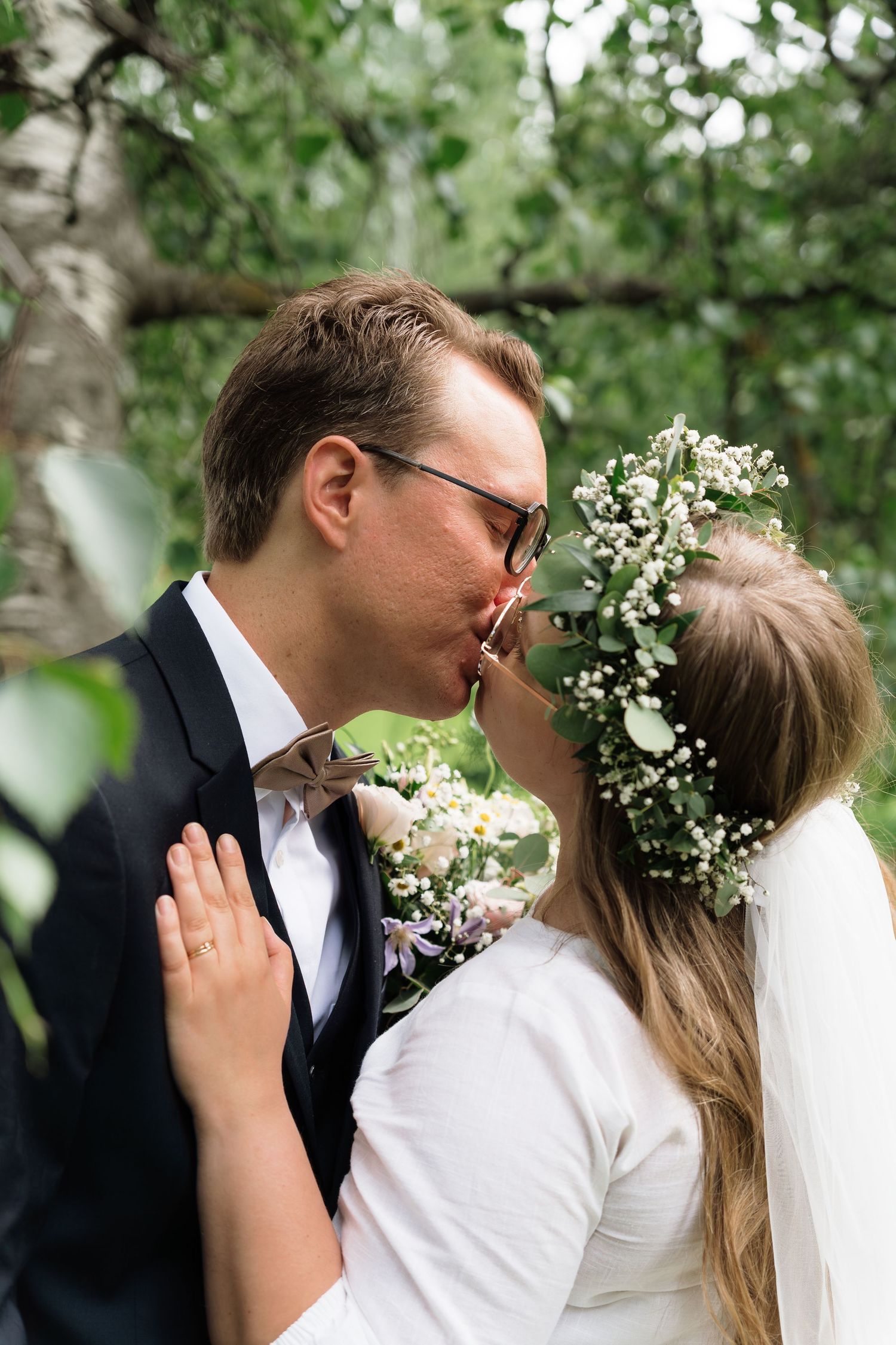 A bride and groom share a kiss outdoors, bride wearing a floral crown with greenery and white flowers on their wedding day.