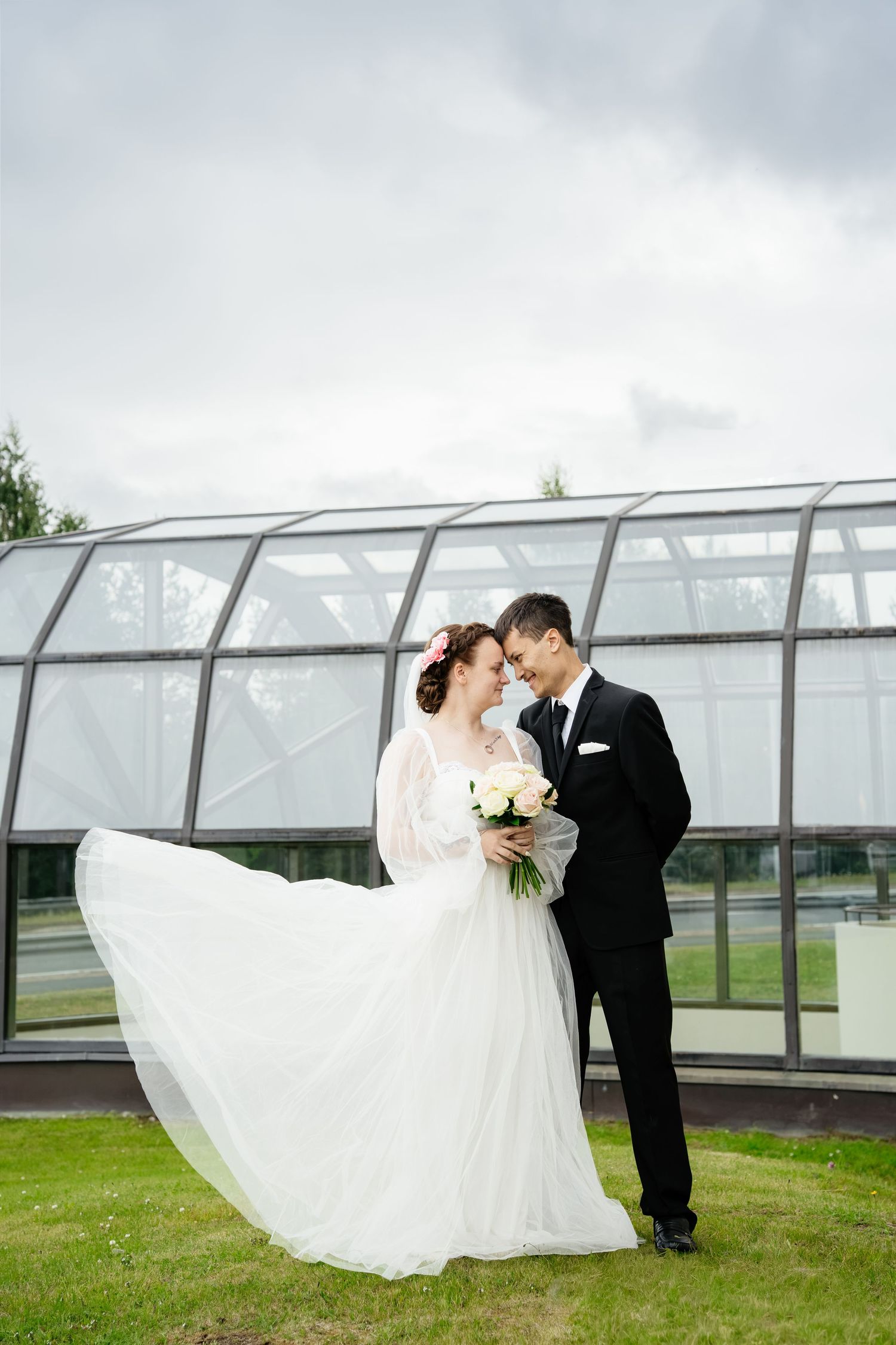 Bride in flowing white dress holding bouquet stands with groom in black suit outdoors near glass building on their wedding day.