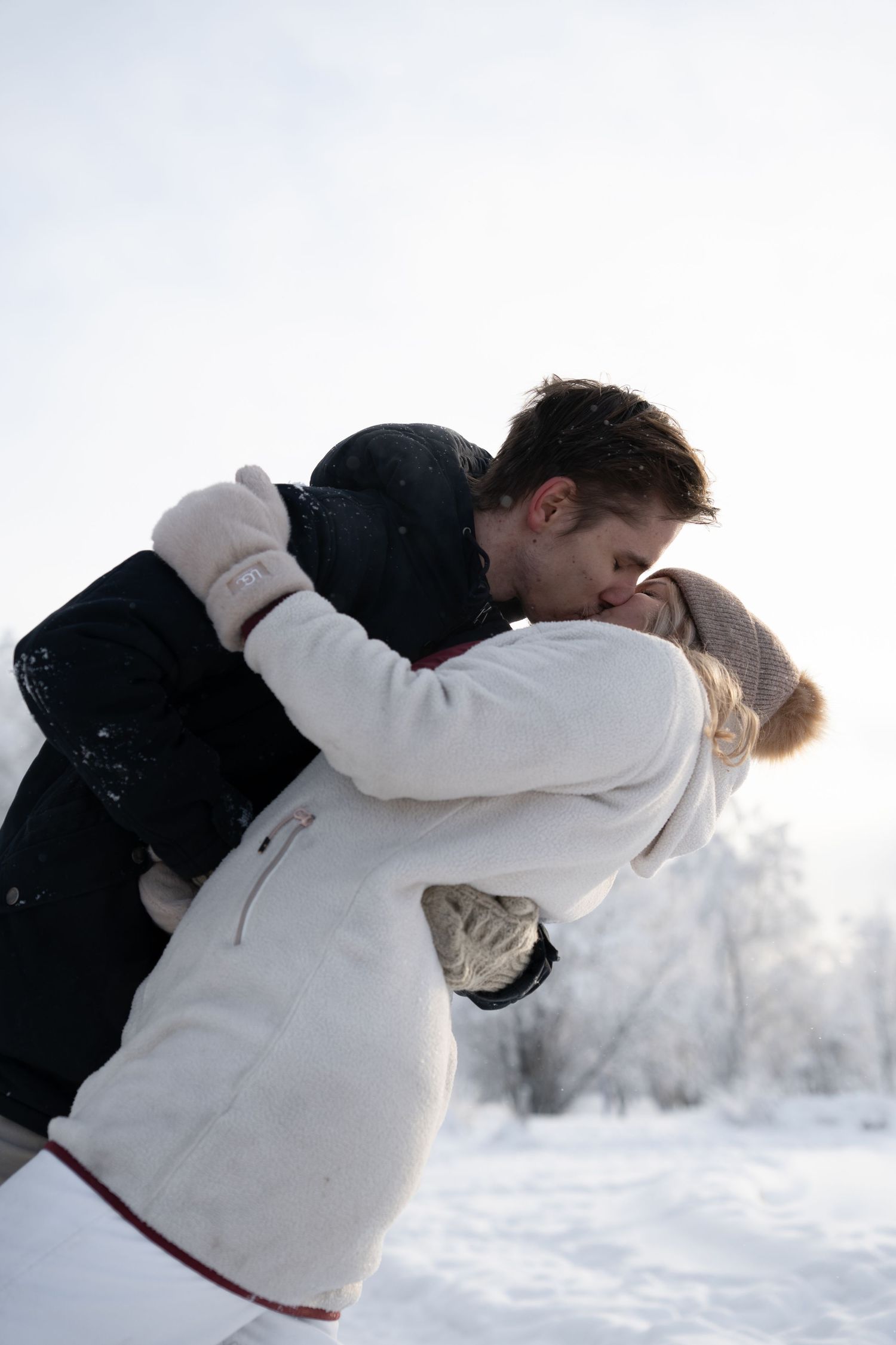 Couple sharing a romantic kiss outdoors in a snowy winter landscape, wearing warm coats and mittens.