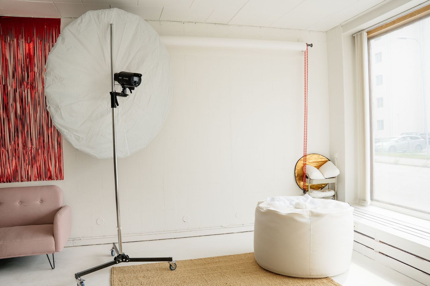 A bright photography studio with a large umbrella light, white backdrop, pink sofa, and modern white ottoman near a window.