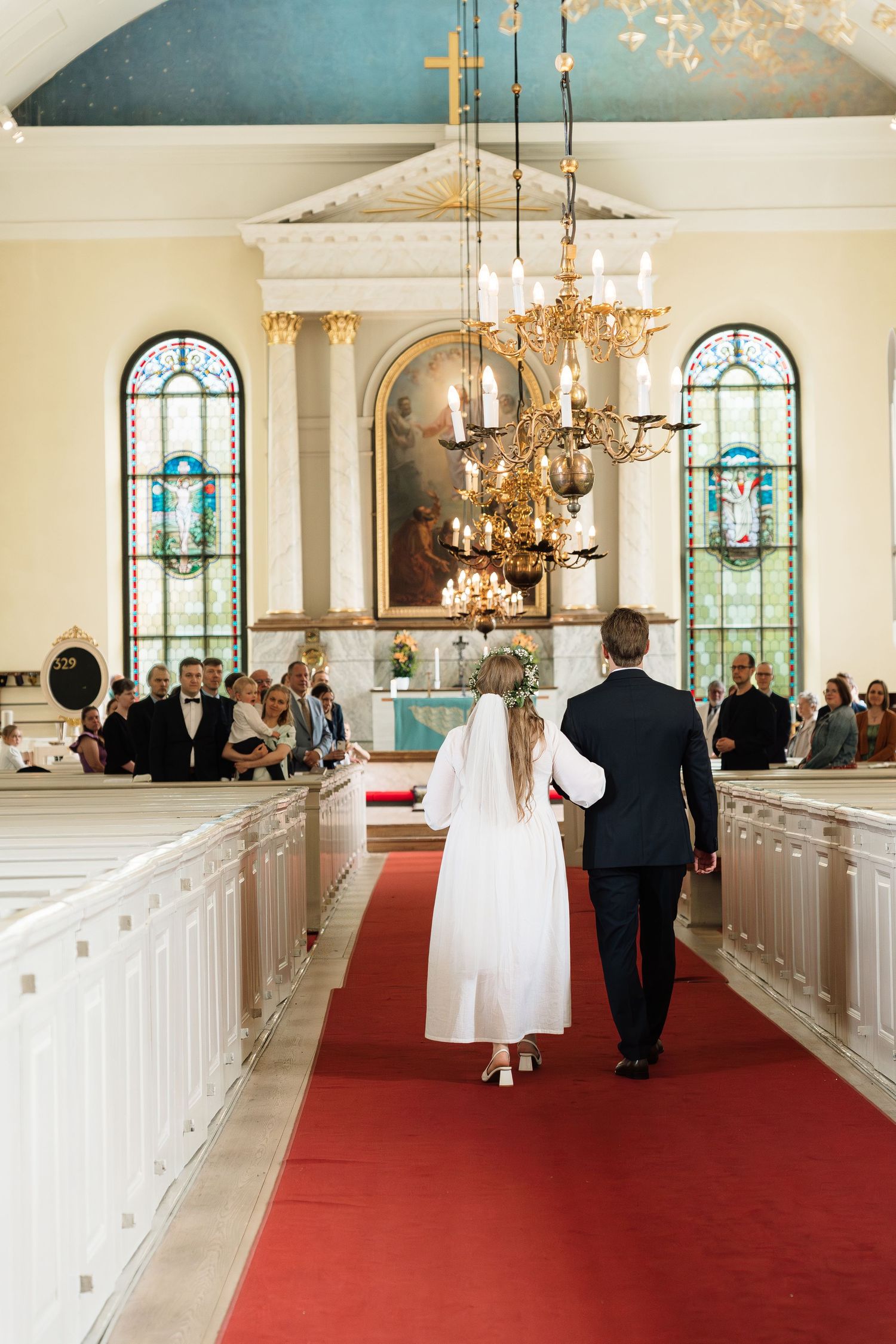 Bride and groom walking down red carpet aisle in elegant white church with stained glass windows and gold chandelier.