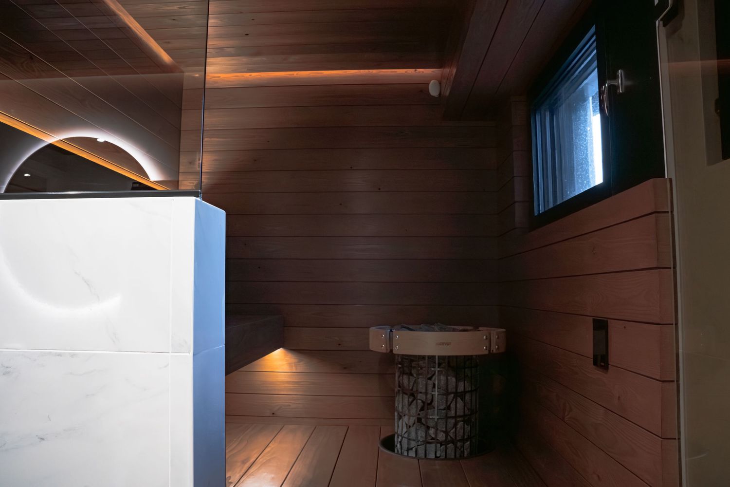 Modern sauna interior with wooden walls, bench, stone heater, and ambient lighting creating a warm relaxing atmosphere.