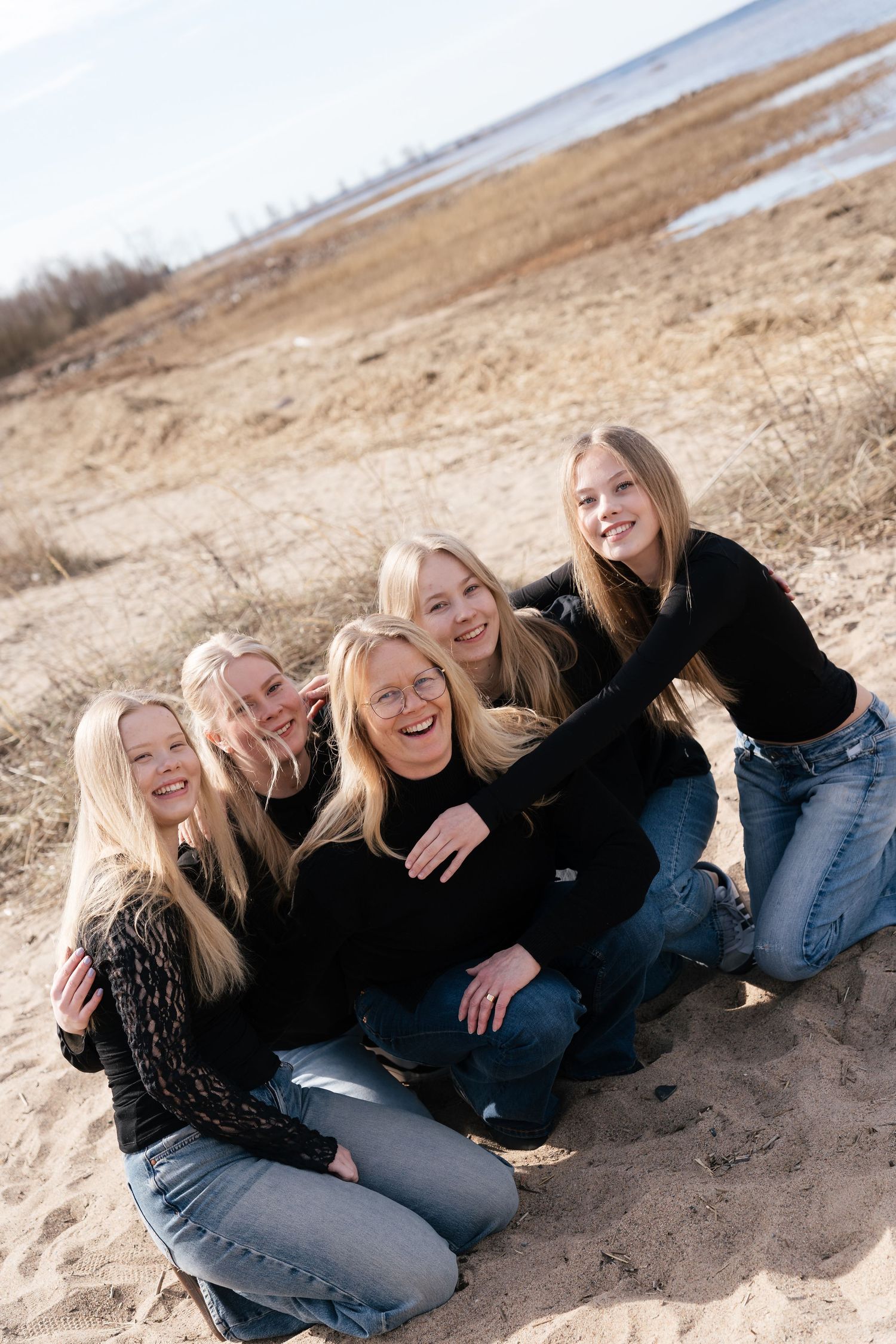 Five blonde women in black tops smile together on a sandy beach on a sunny day.