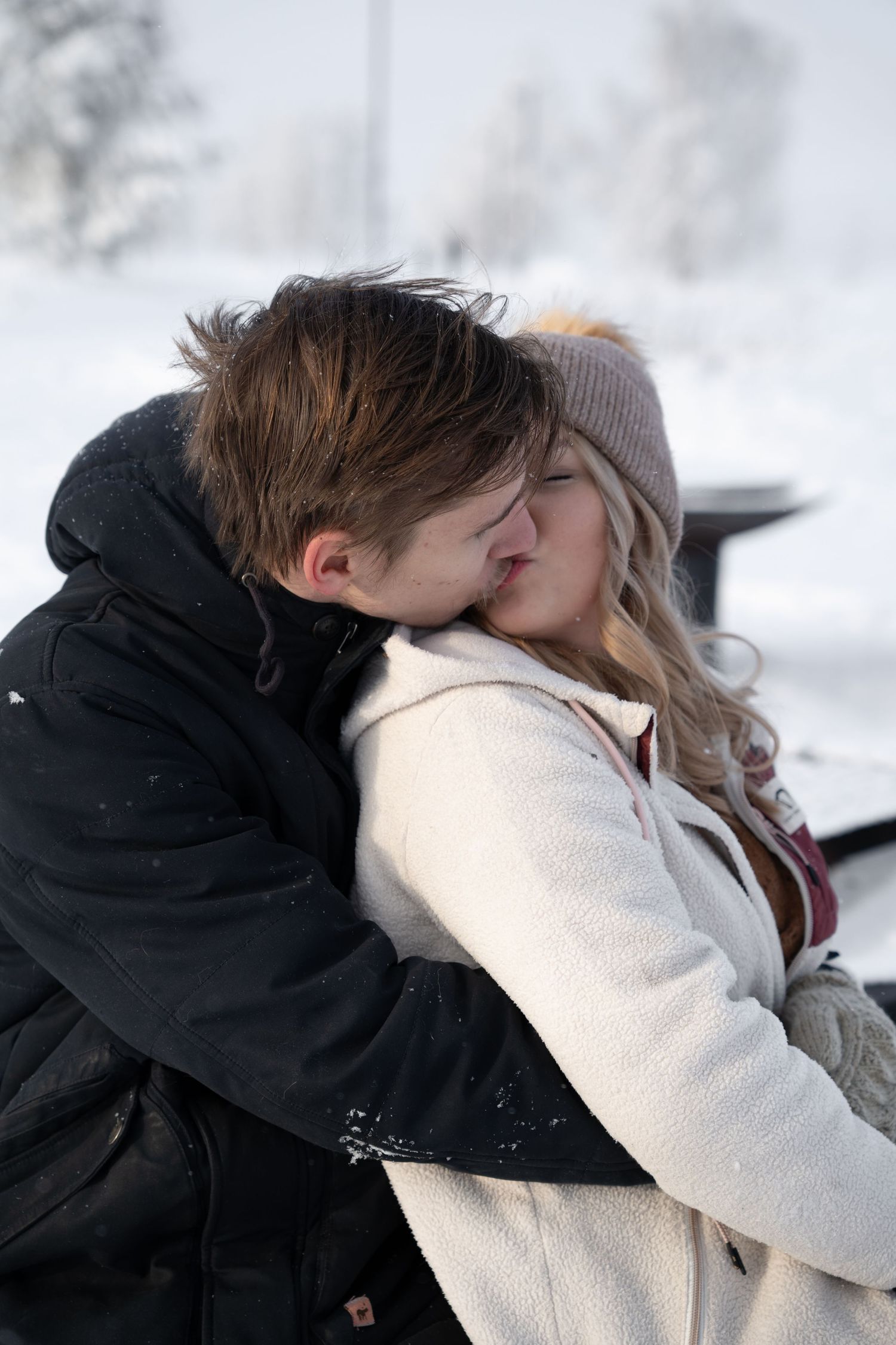 Couple sharing a tender kiss while seated outdoors in winter, surrounded by snow-covered trees, woman in white coat and beanie.