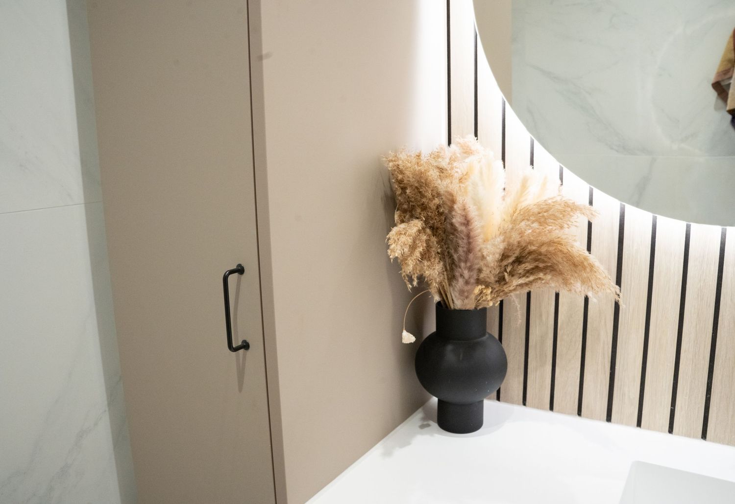 Black vase with dried pampas grass on white countertop beside beige cabinet and round mirror in modern bathroom.