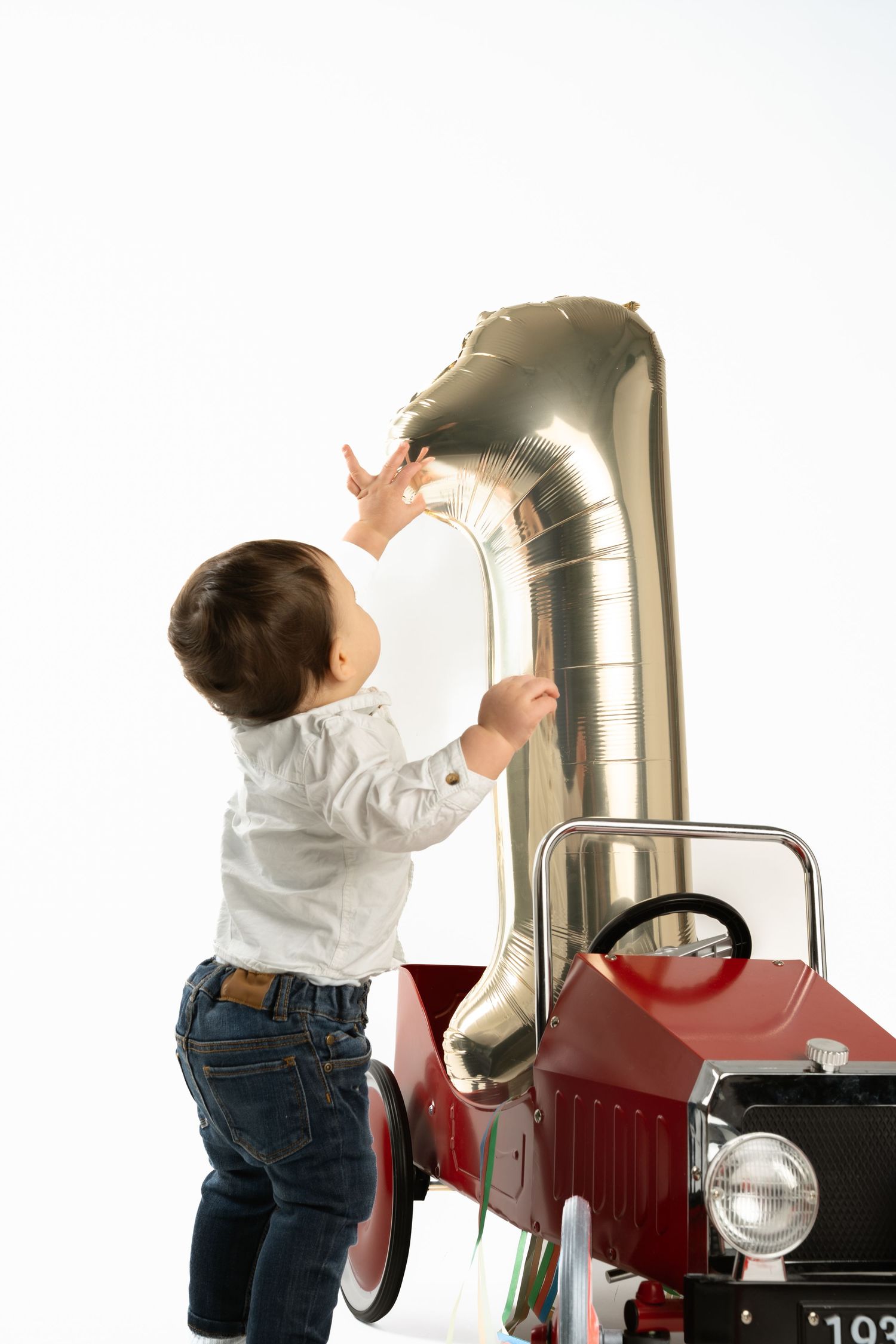 Toddler boy in jeans reaching up to touch a silver number one balloon beside a red vintage pedal car.