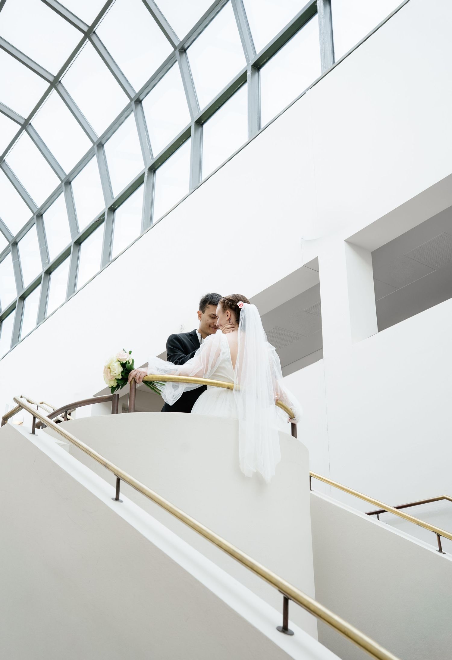 Bride and groom share a romantic moment on a grand staircase with gold railings in a modern white venue.