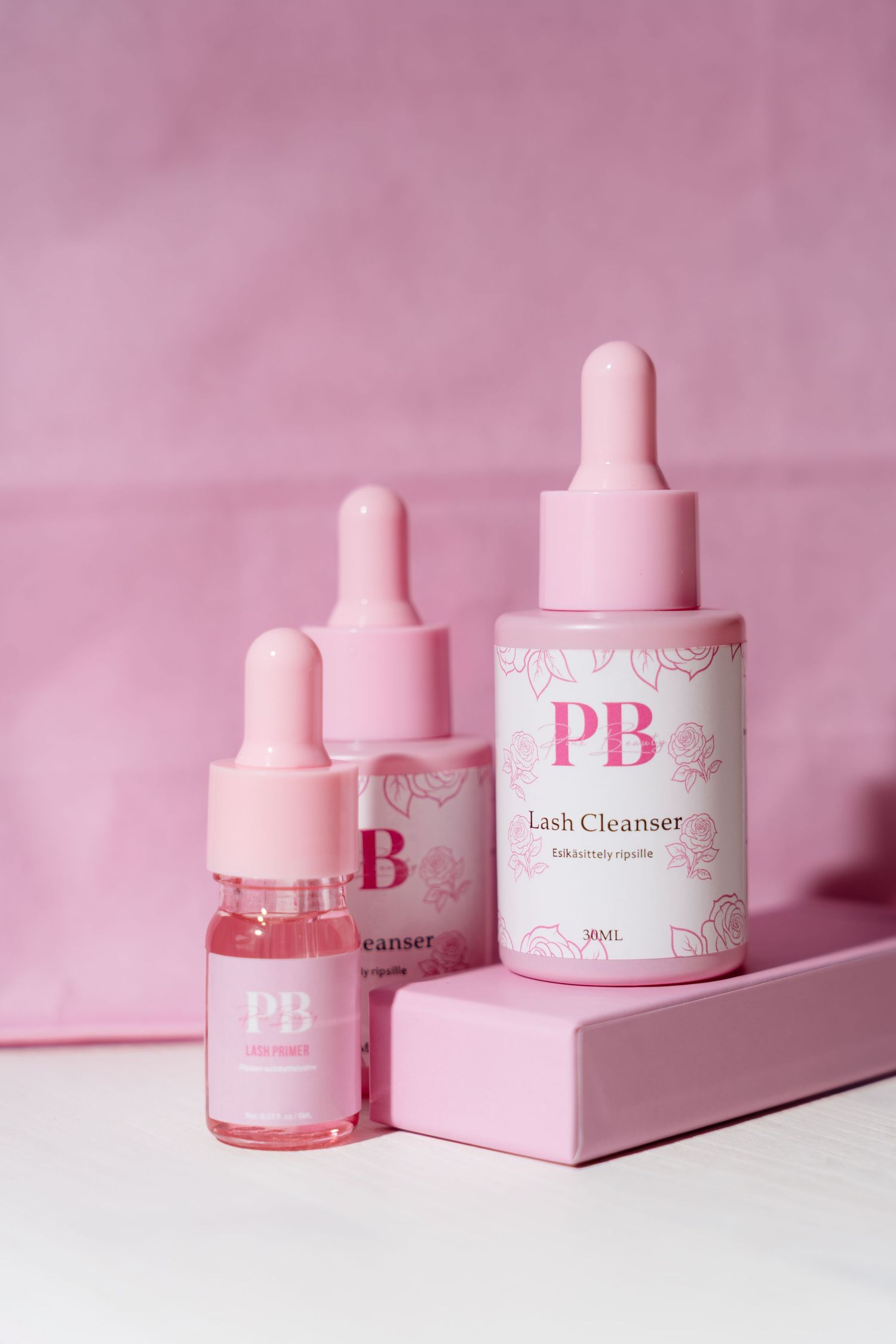 Three pink PB Lash Cleanser dropper bottles arranged on pink geometric risers.