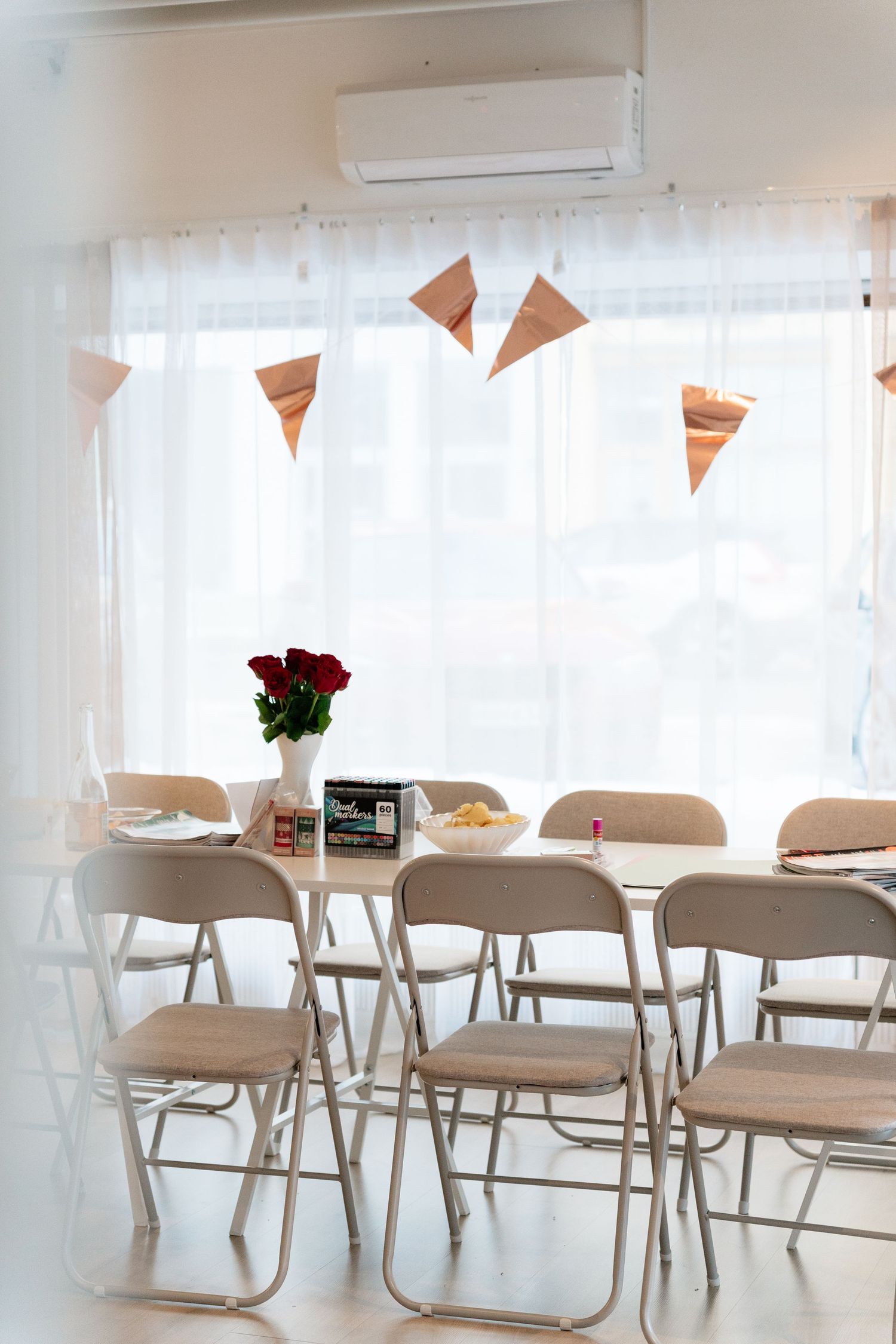 Bright party room with folding chairs, long table, red roses, snacks, and rose gold triangle bunting near a window.