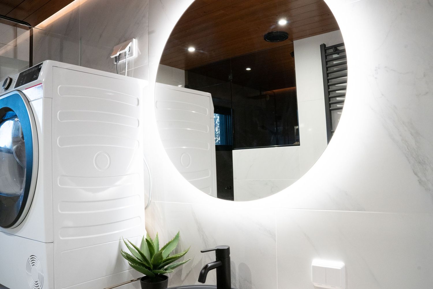Modern bathroom with round LED mirror, white marble walls, black faucet sink, and stacked washer unit with wood ceiling accents.