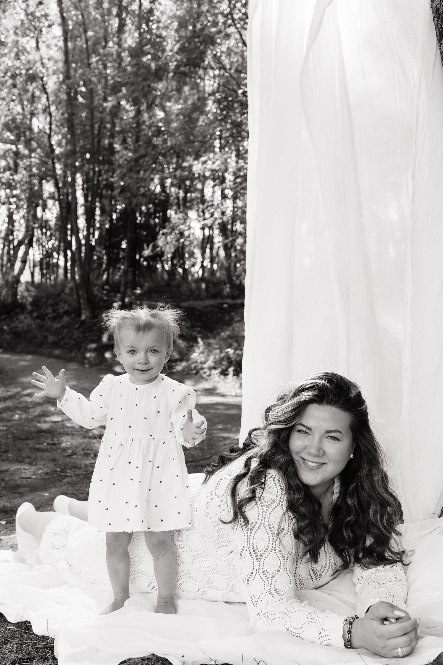 Black and white photo of a smiling mother in white lace dress lying on blanket with toddler daughter outdoors.