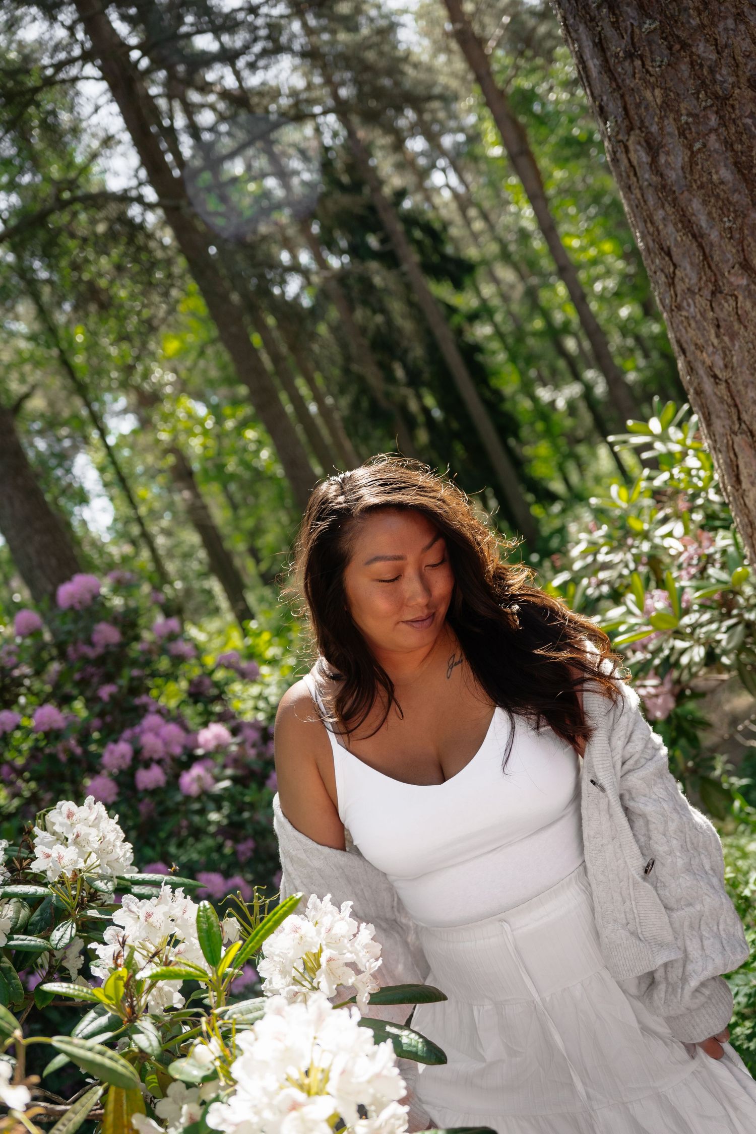 Woman in white dress admiring blooming white flowers in a lush green forest garden on a sunny day.
