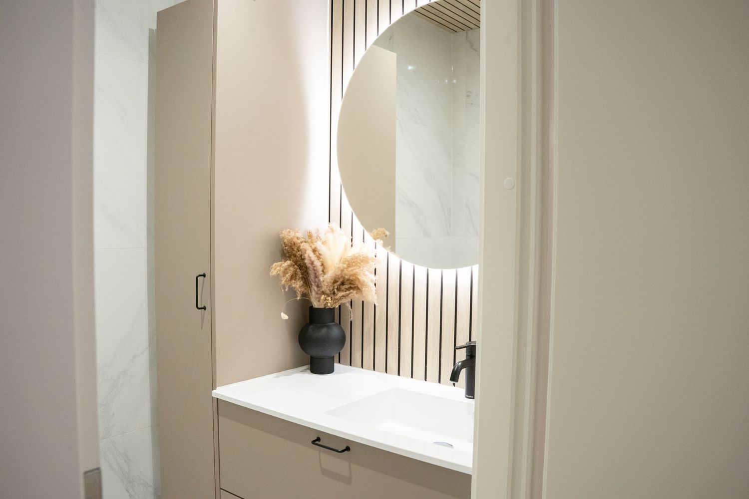 Modern bathroom vanity with round backlit mirror, white cabinet, black faucet, and decorative pampas grass in black vase.