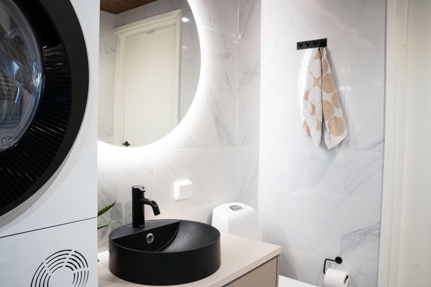 Modern bathroom with black round vessel sink, illuminated round mirror, marble walls, and washing machine.