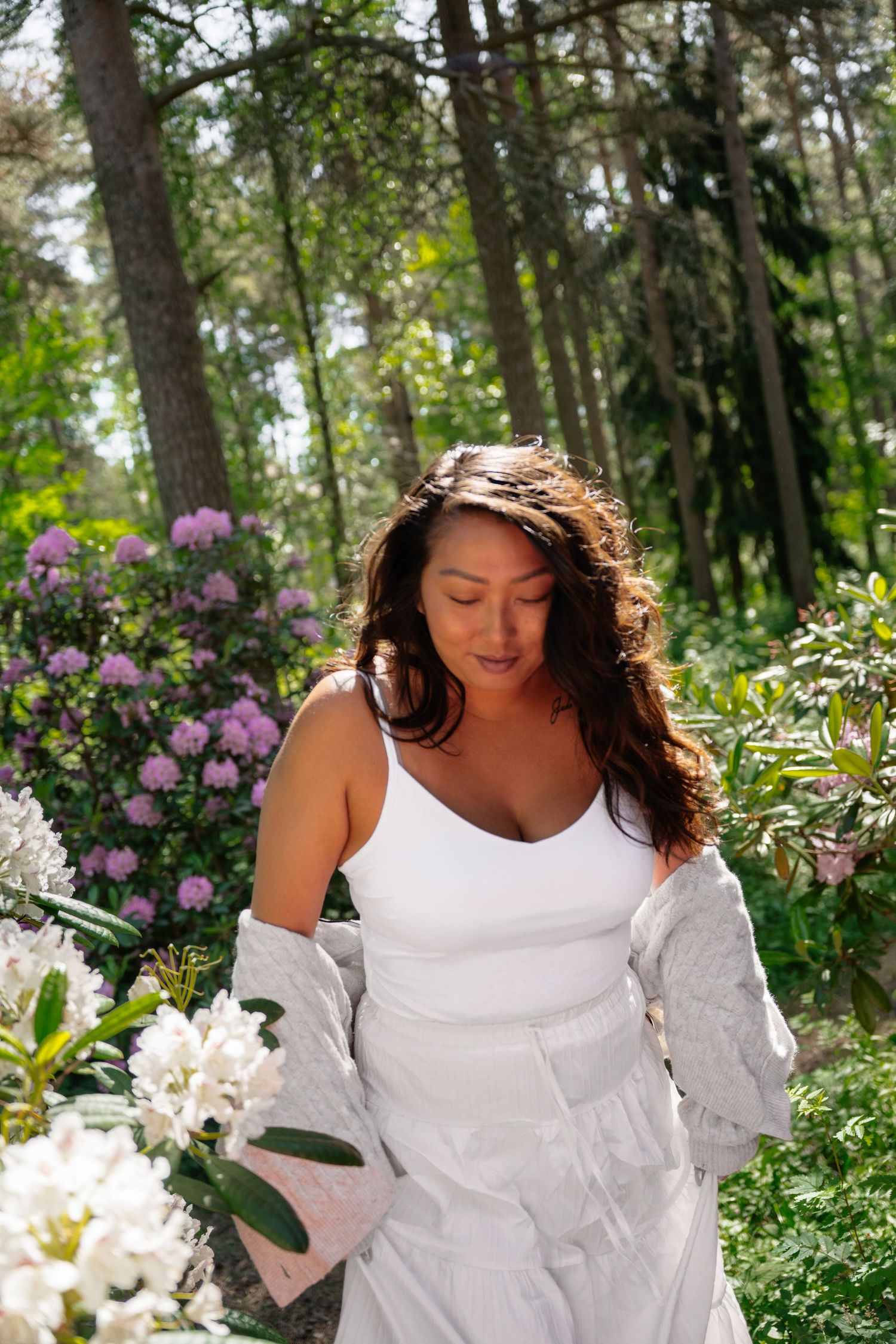 Woman in white dress and jacket among blooming rhododendrons in a lush green forest garden.