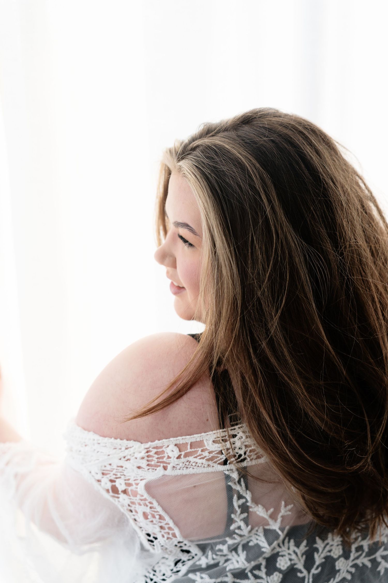 Young brunette woman in lace lingerie posing in soft backlit white light, side profile view.