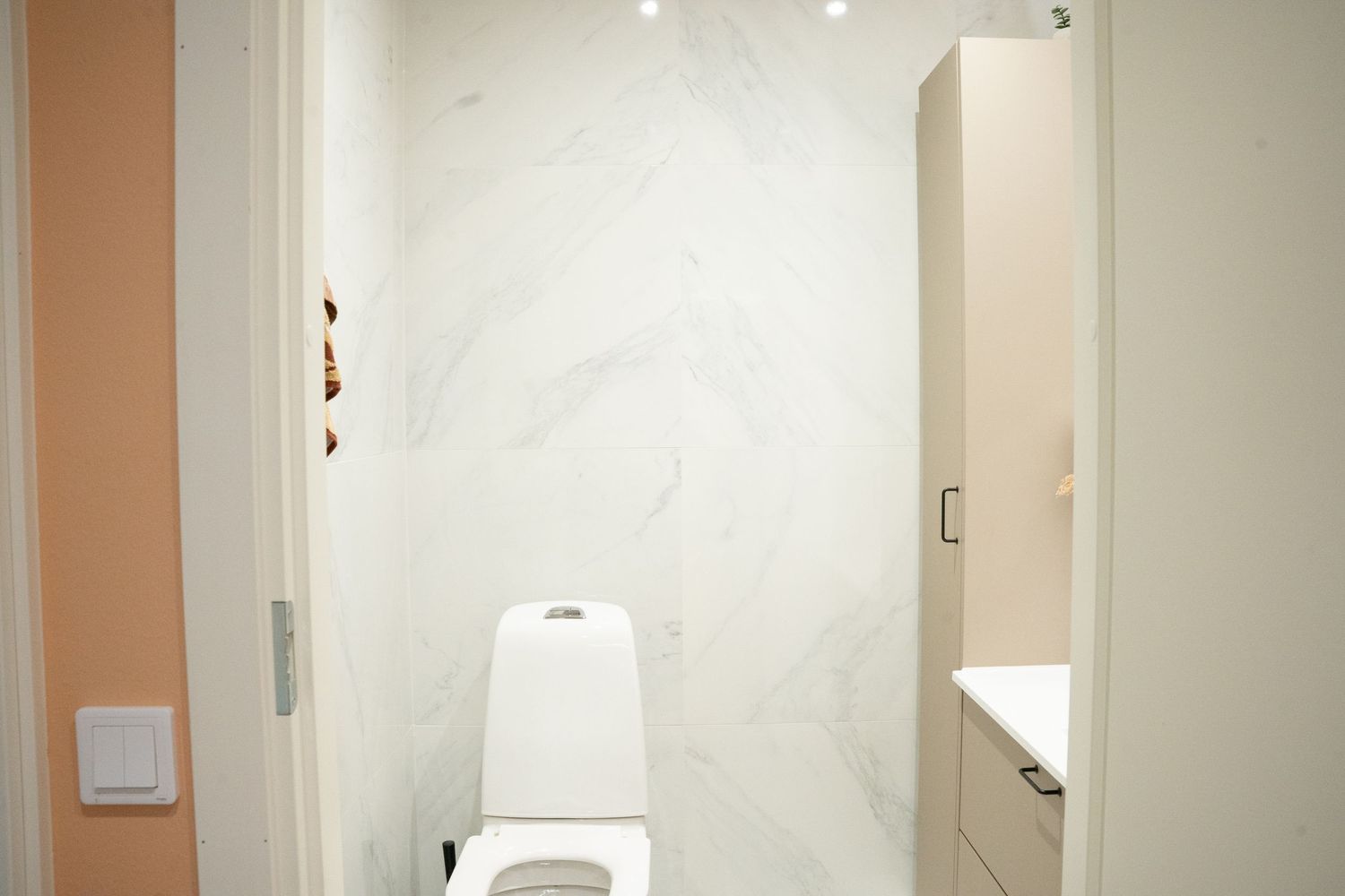 Modern white bathroom with marble wall tiles, white toilet, and built-in storage cabinet in a minimalist design.