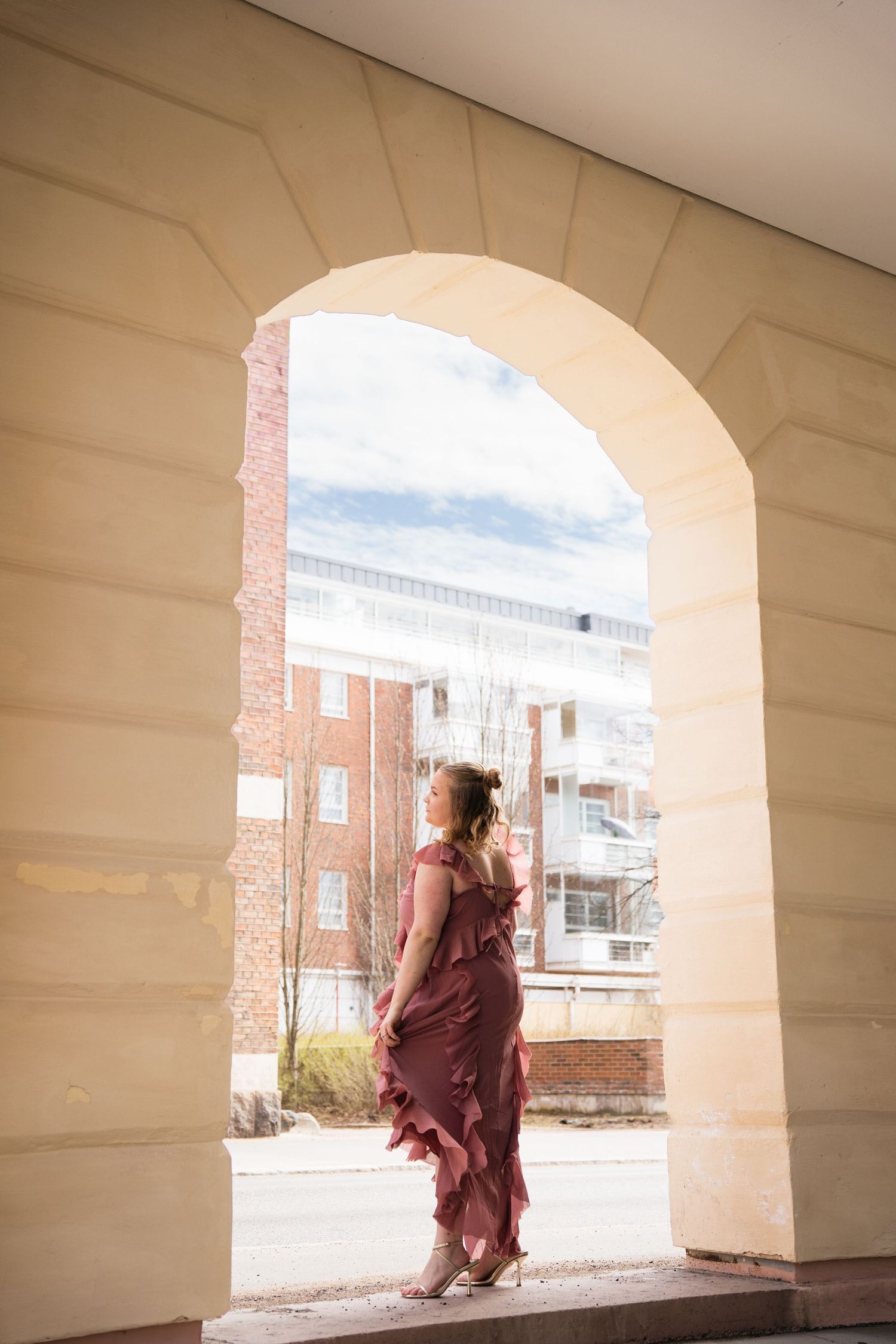 Woman in a flowing burgundy ruffle dress standing in an elegant stone archway with urban buildings in the background.