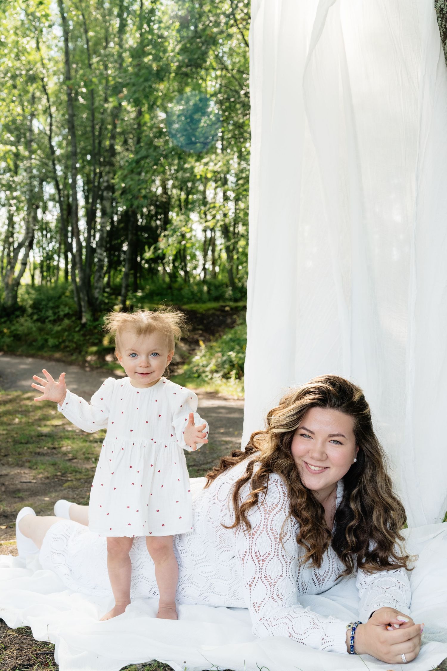 Mother in white lace dress lying on blanket outdoors smiling at toddler girl in white dress standing in sunlit forest.