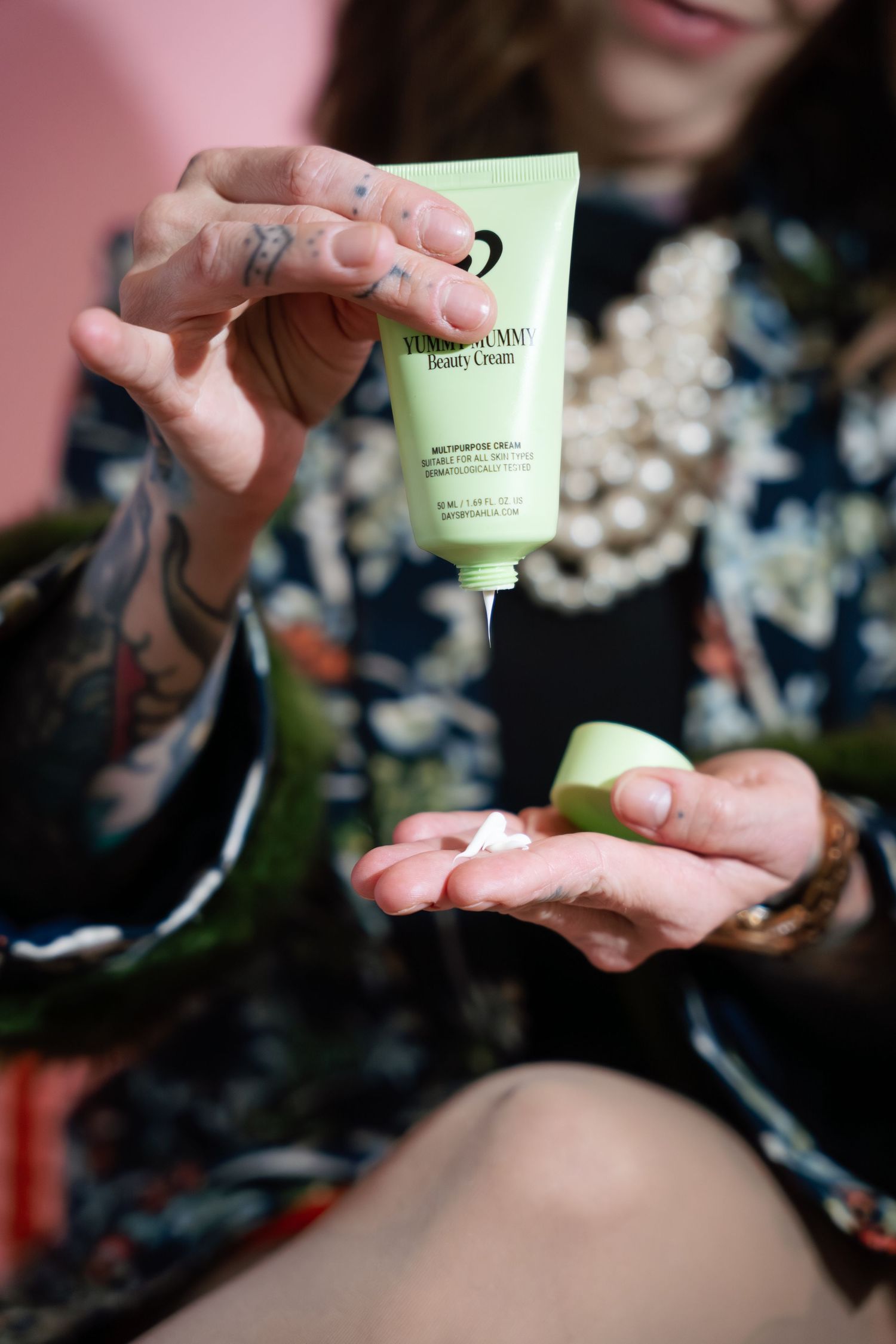 Tattooed person squeezing Days by Dahlia Yummy Mummy Beauty Cream from green tube into palm.