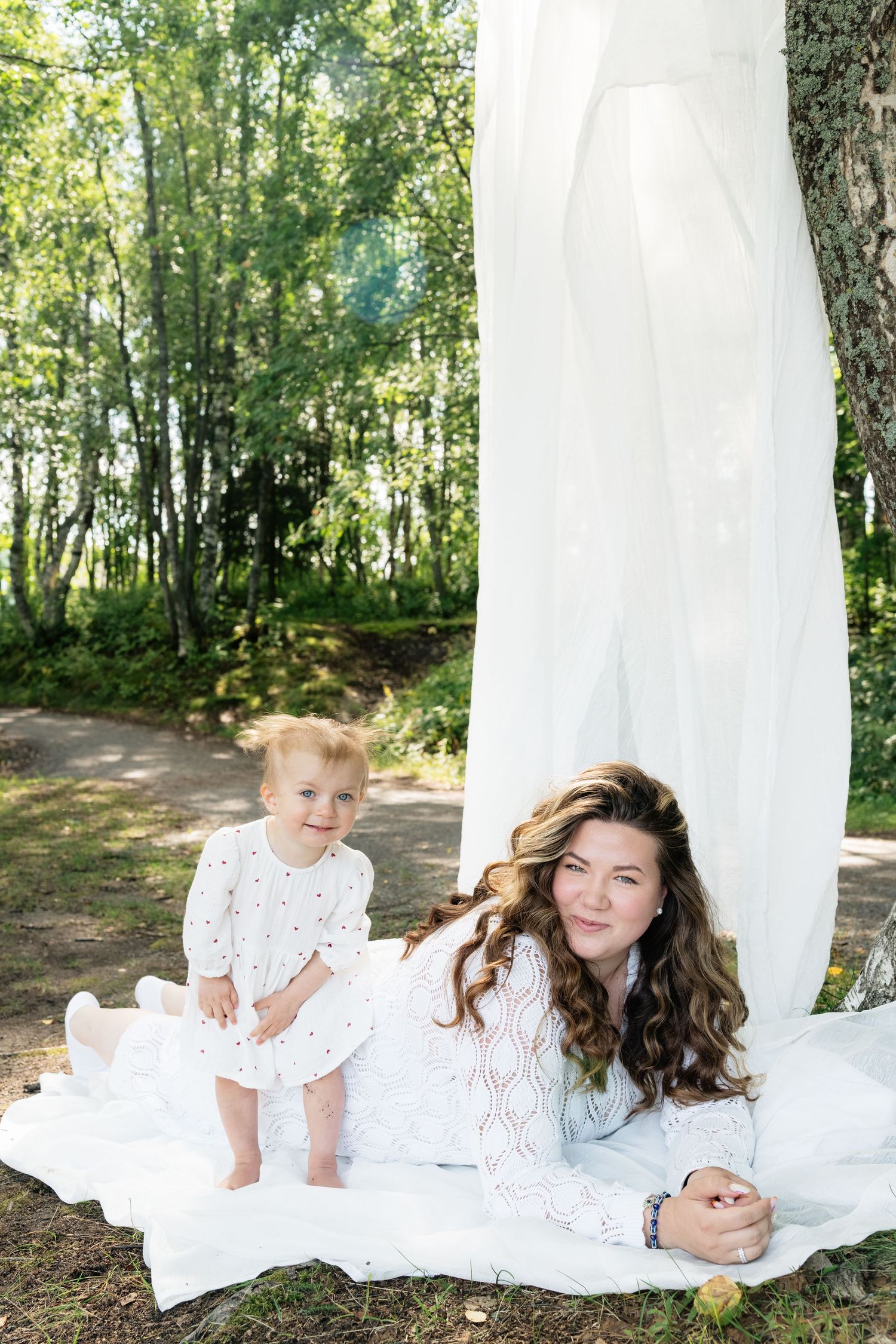 Mother in white lace dress lying outdoors with smiling toddler girl in forest setting.