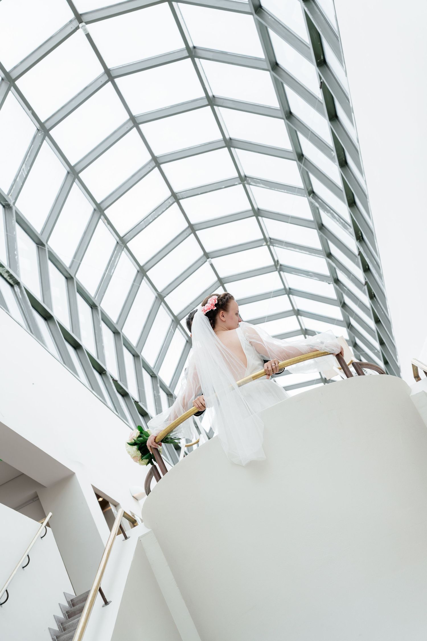 Bride in white gown and veil stands on curved staircase under glass atrium ceiling, holding bouquet.