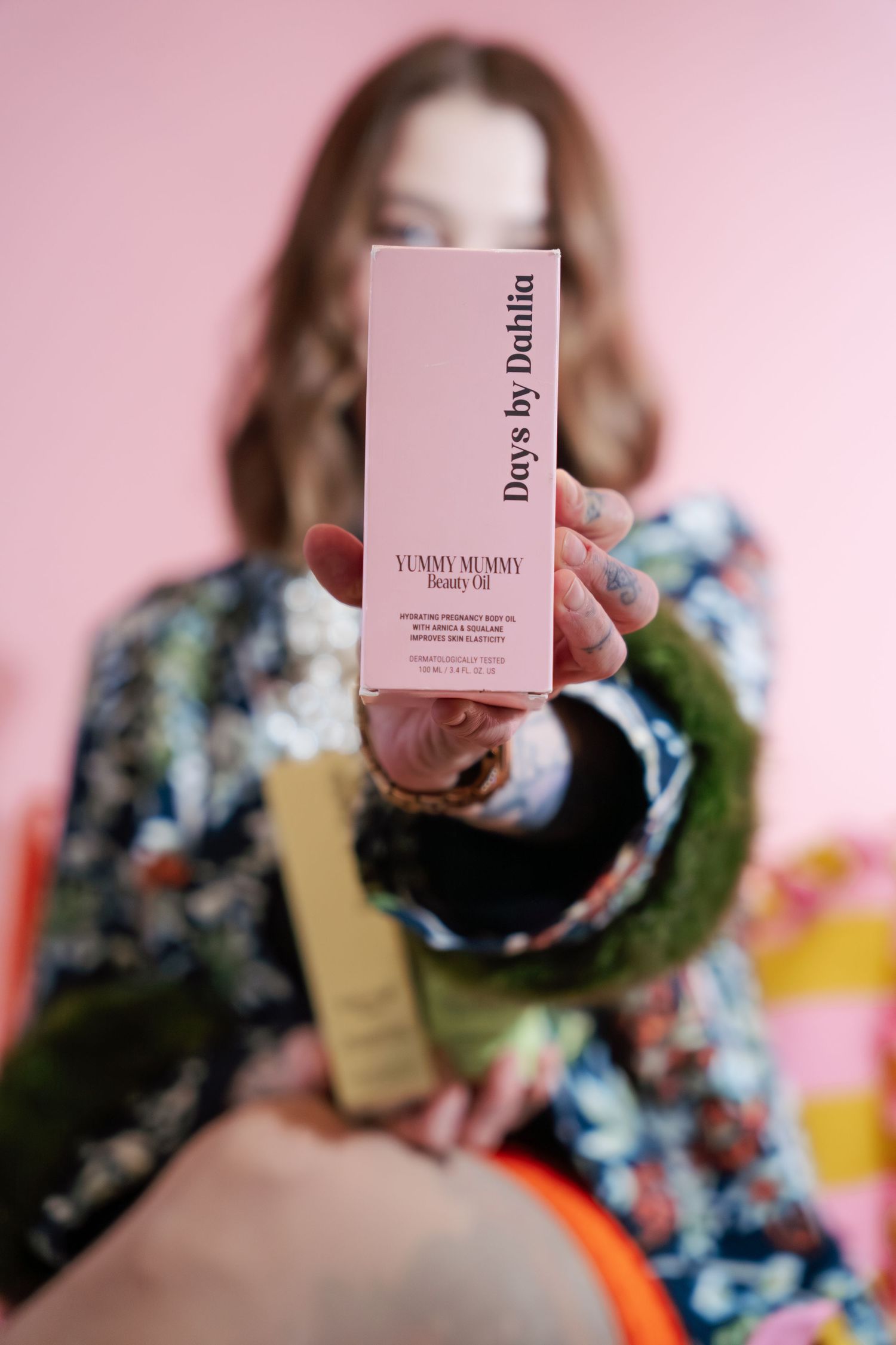 Woman holding Days by Dahlia Yummy Mummy Beauty Oil box toward camera against pink backdrop.