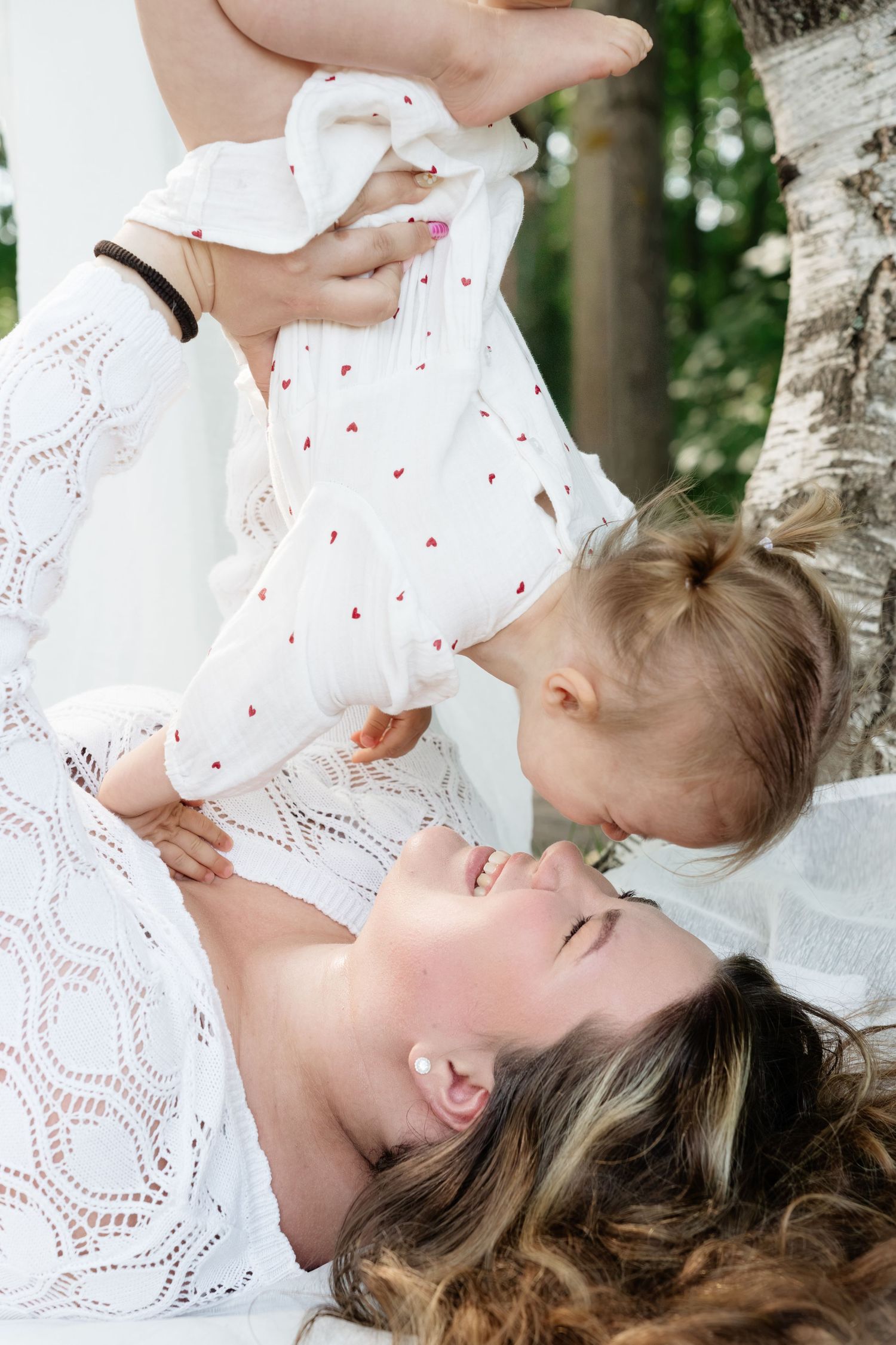 A joyful mother lying down lifts her smiling toddler daughter in a sunny outdoor setting, both wearing white dresses.