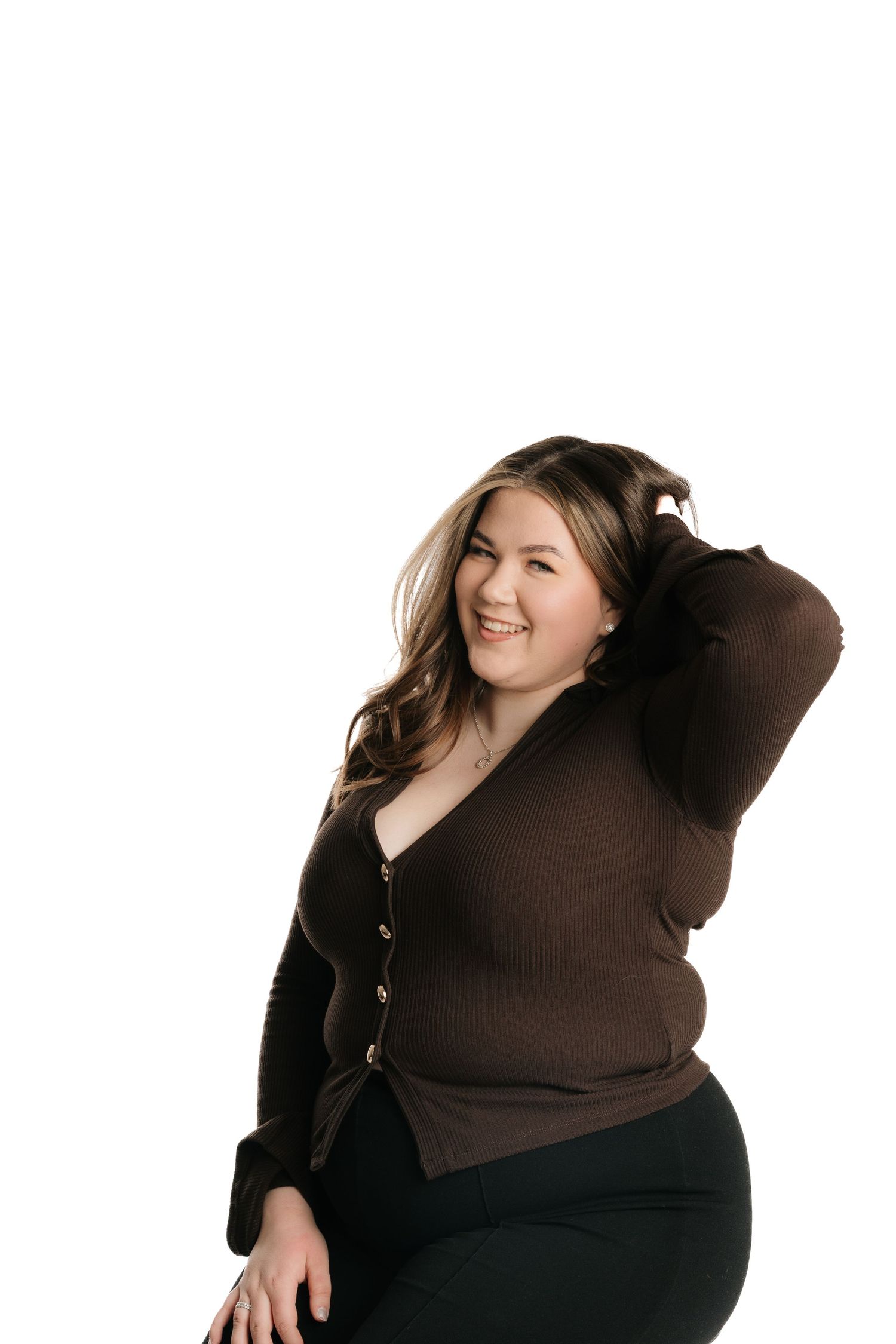 Smiling plus-size woman in brown cardigan and black pants posing with hand in hair against white background.