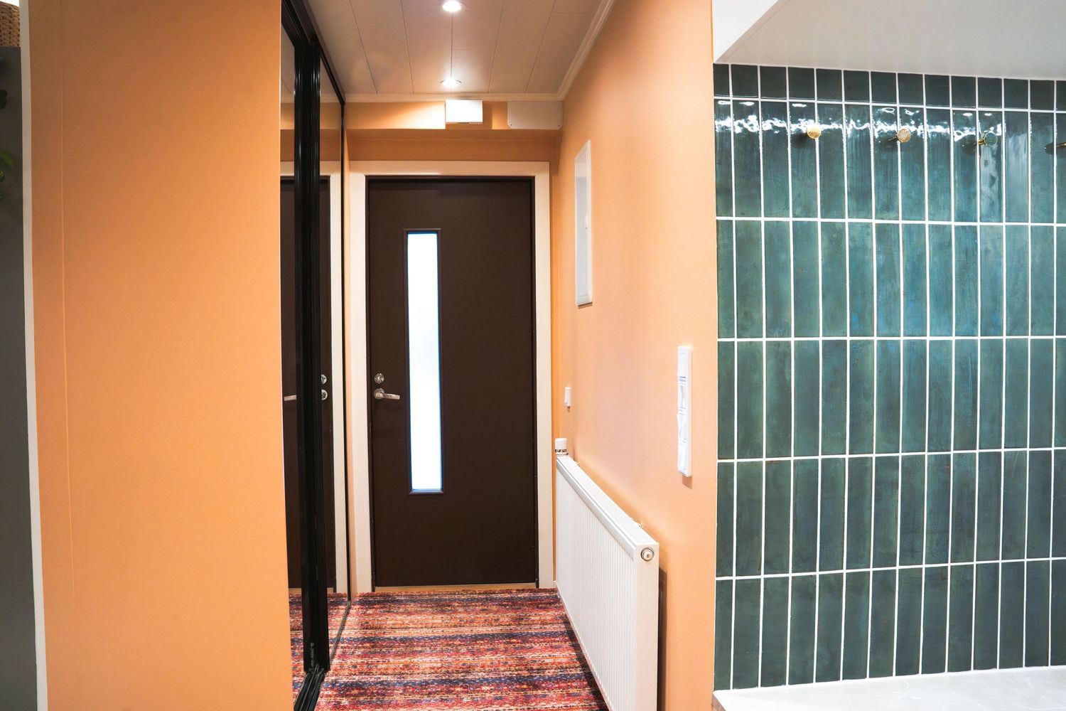 Narrow hallway with orange walls, dark wood door, patterned rug, and green tiled bathroom entrance with white radiator.