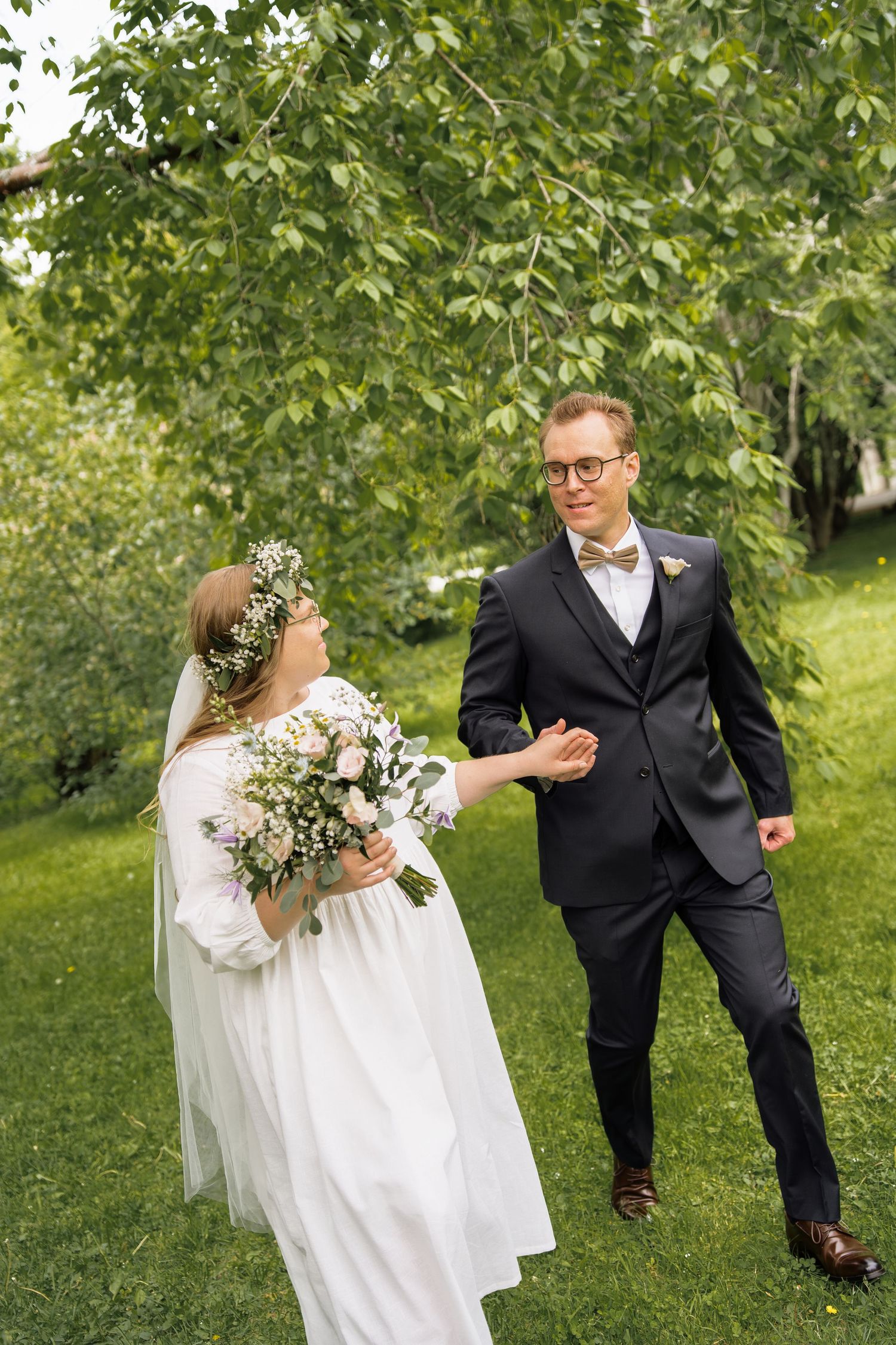Bride in white dress and floral crown holds bouquet while groom in dark suit smiles at her in lush green garden.