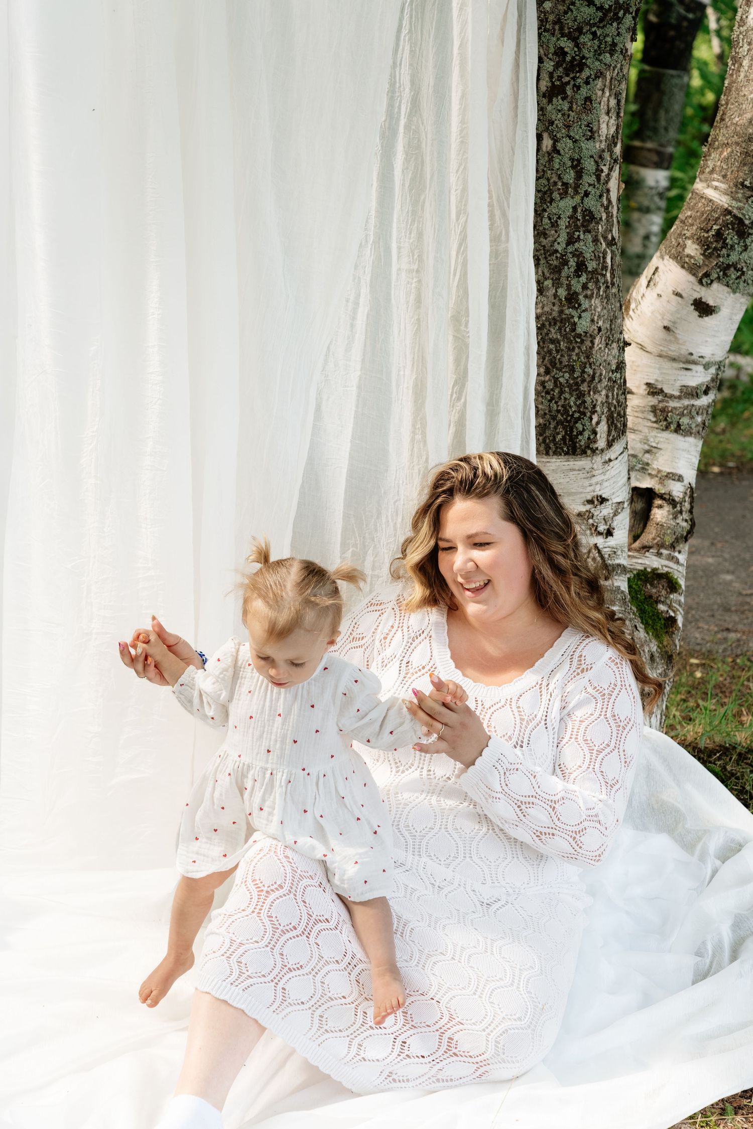 Smiling mother in white lace dress sits on blanket with toddler daughter near birch trees and white curtain outdoors.