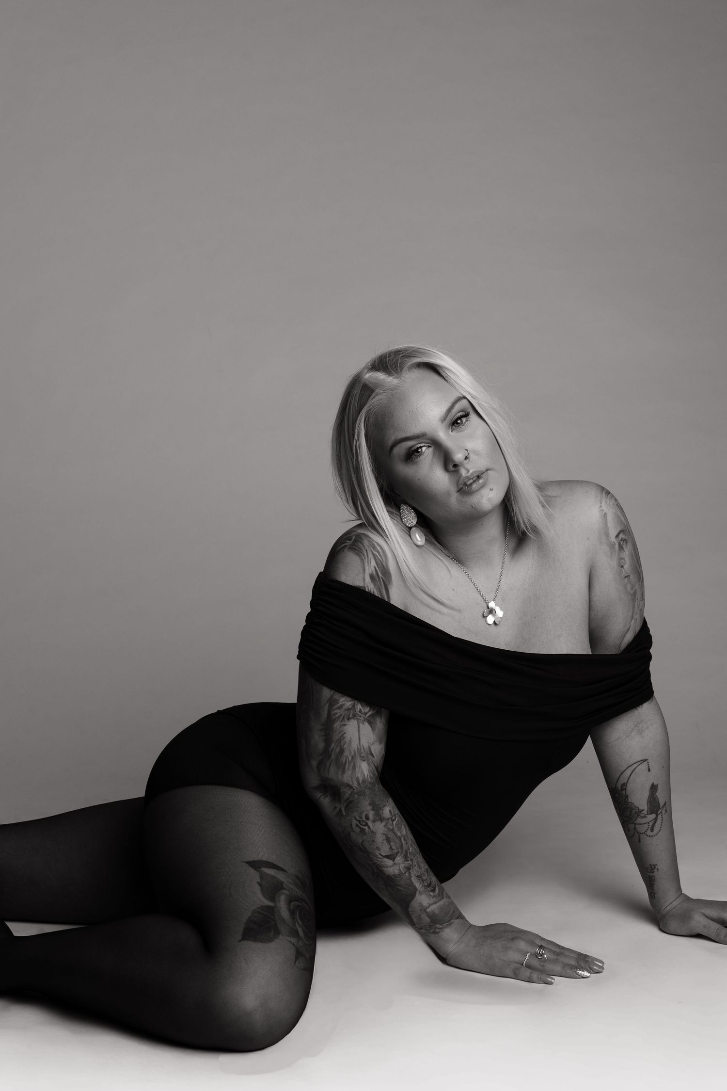 Black and white portrait of tattooed blonde woman in black off-shoulder bodysuit posing on floor.