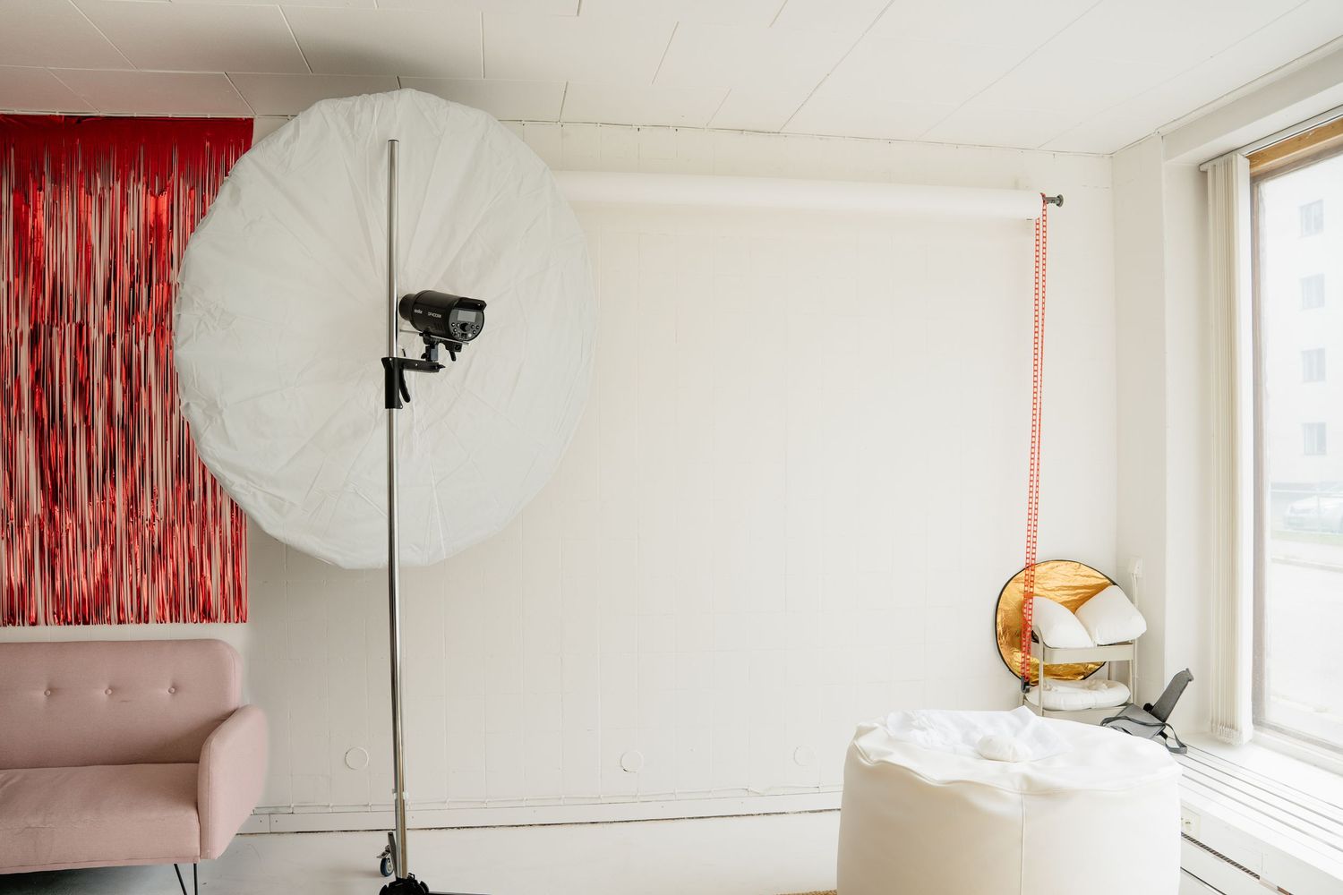 Professional photo studio with large white umbrella light, red backdrop, pink sofa, and natural window light.