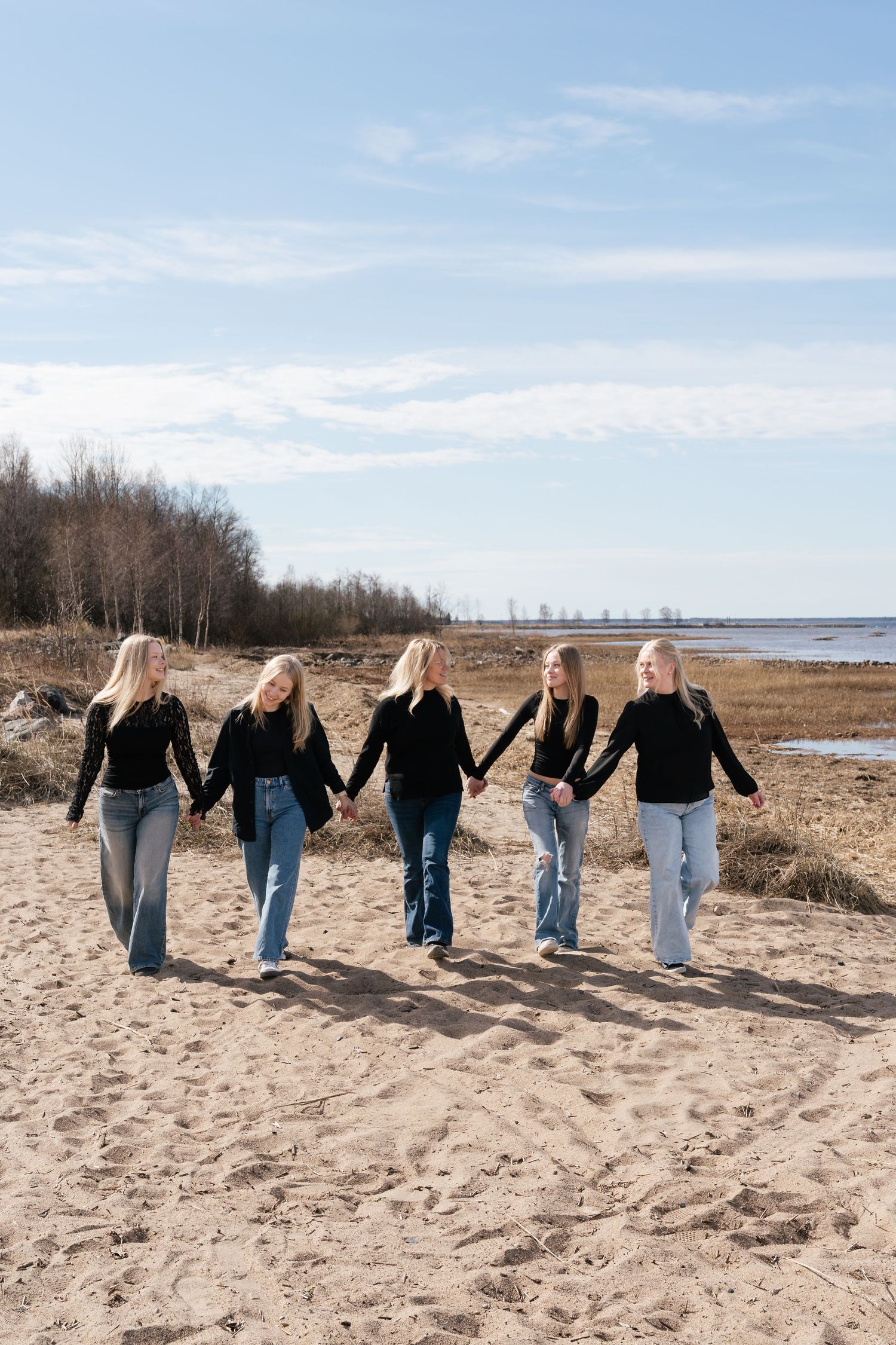 Five women in black tops and jeans walk hand in hand along a sandy beach under a cloudy sky.