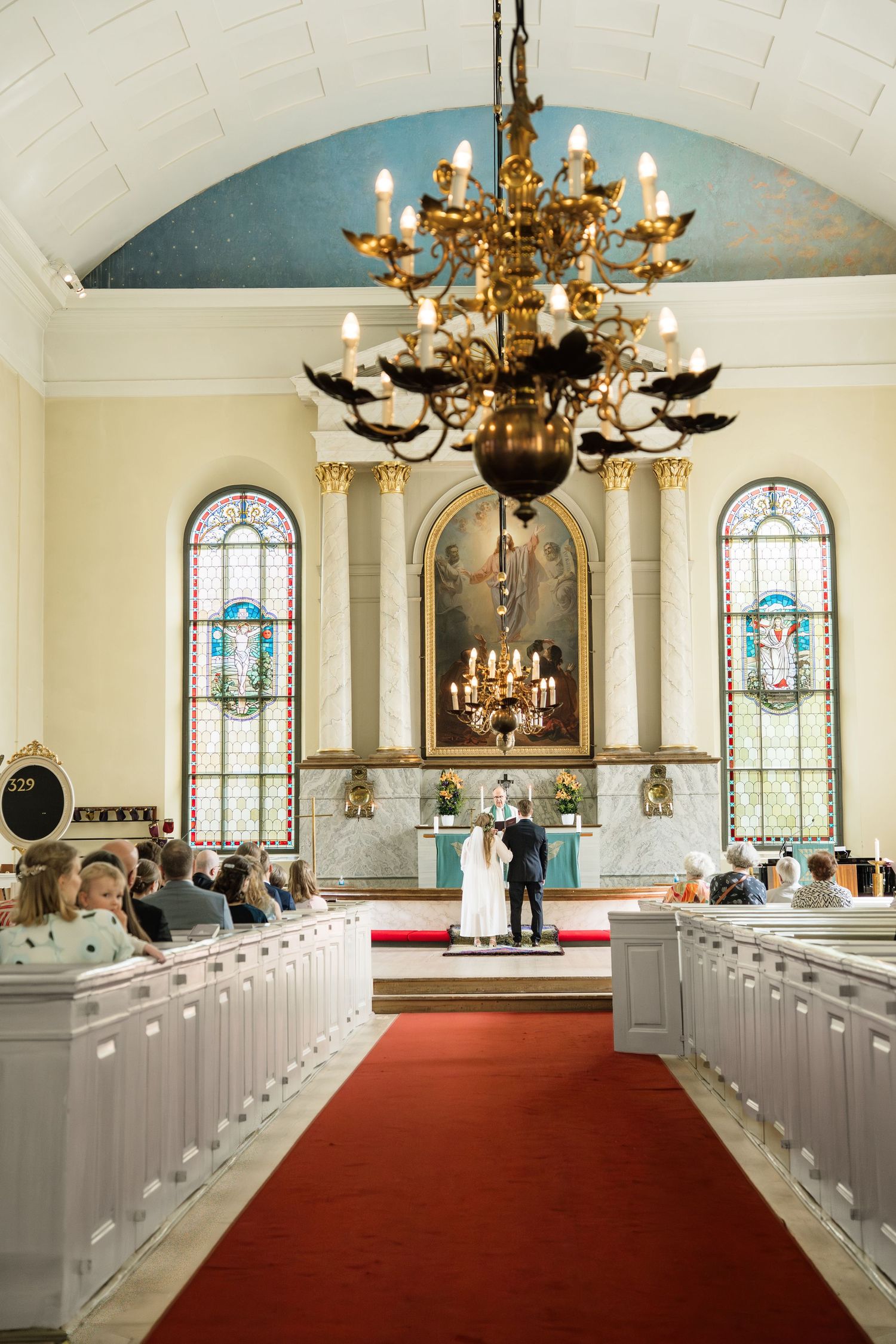 Bride and groom stand at the altar of an elegant church with stained glass windows, red carpet aisle, and golden chandelier.
