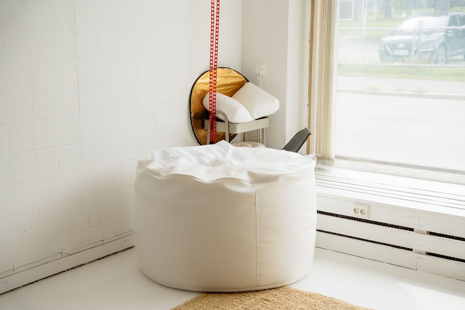 White bean bag chair near a bright window with a round mirror and red cord on a white wall.