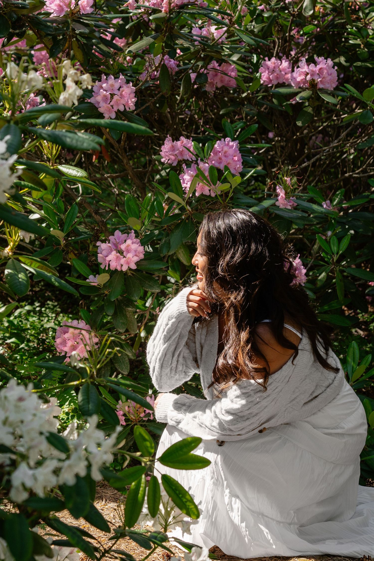 Woman in white dress sitting among blooming pink rhododendron bushes in a sunny garden.