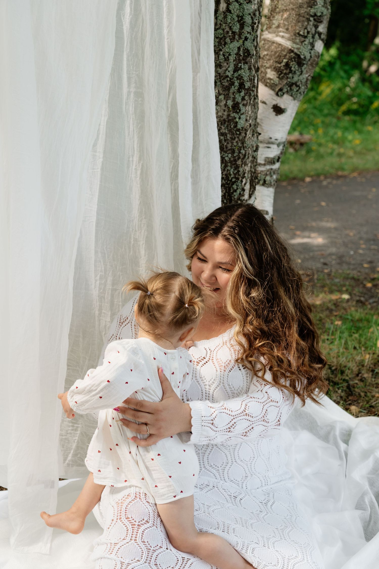 A smiling mother with curly hair in a white lace dress holds her toddler daughter outdoors near white curtains.