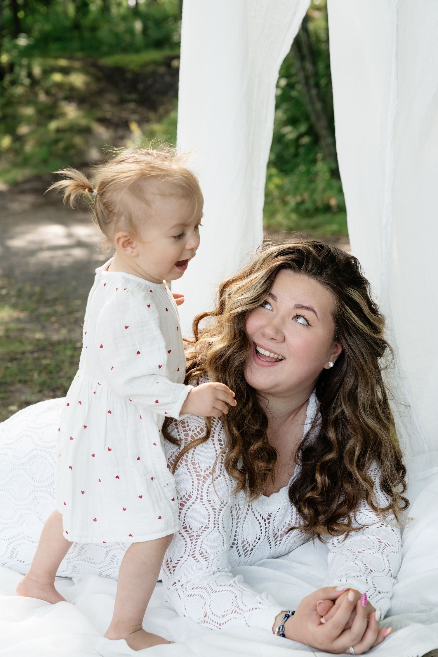 A smiling mother in white dress sits outdoors with her toddler daughter in a white floral dress under a canopy.